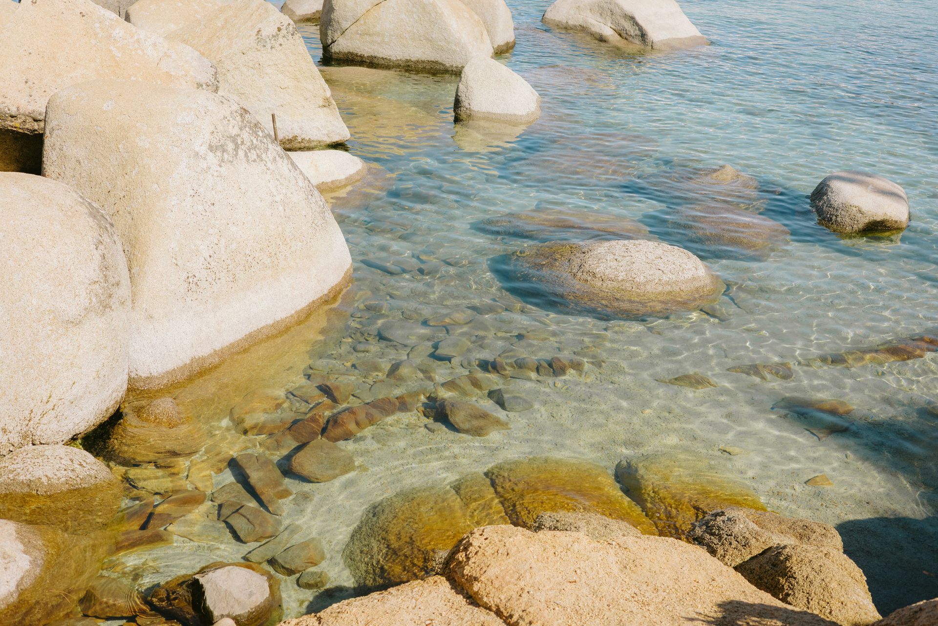 Large, beige boulders in clear, shallow water. Sunlight reflects on the water's surface, revealing the rocky seabed.