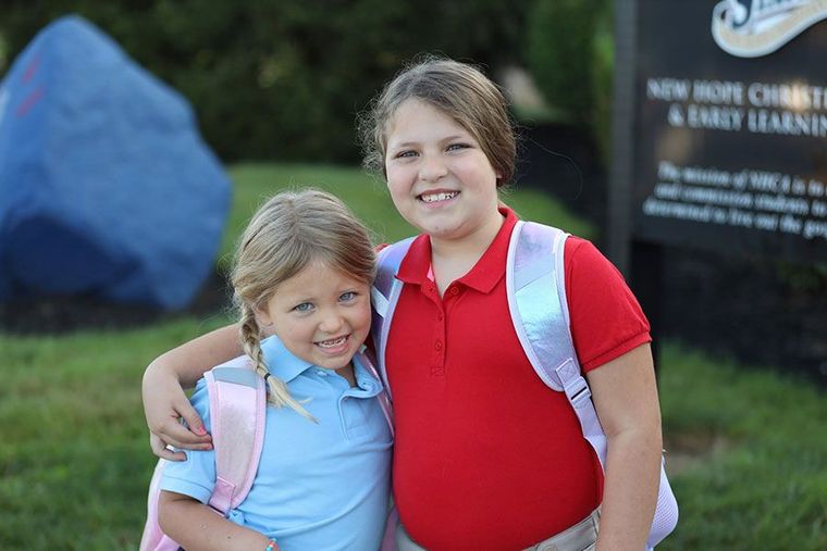 Two children standing outdoors, smiling, with backpacks and colorful shirts