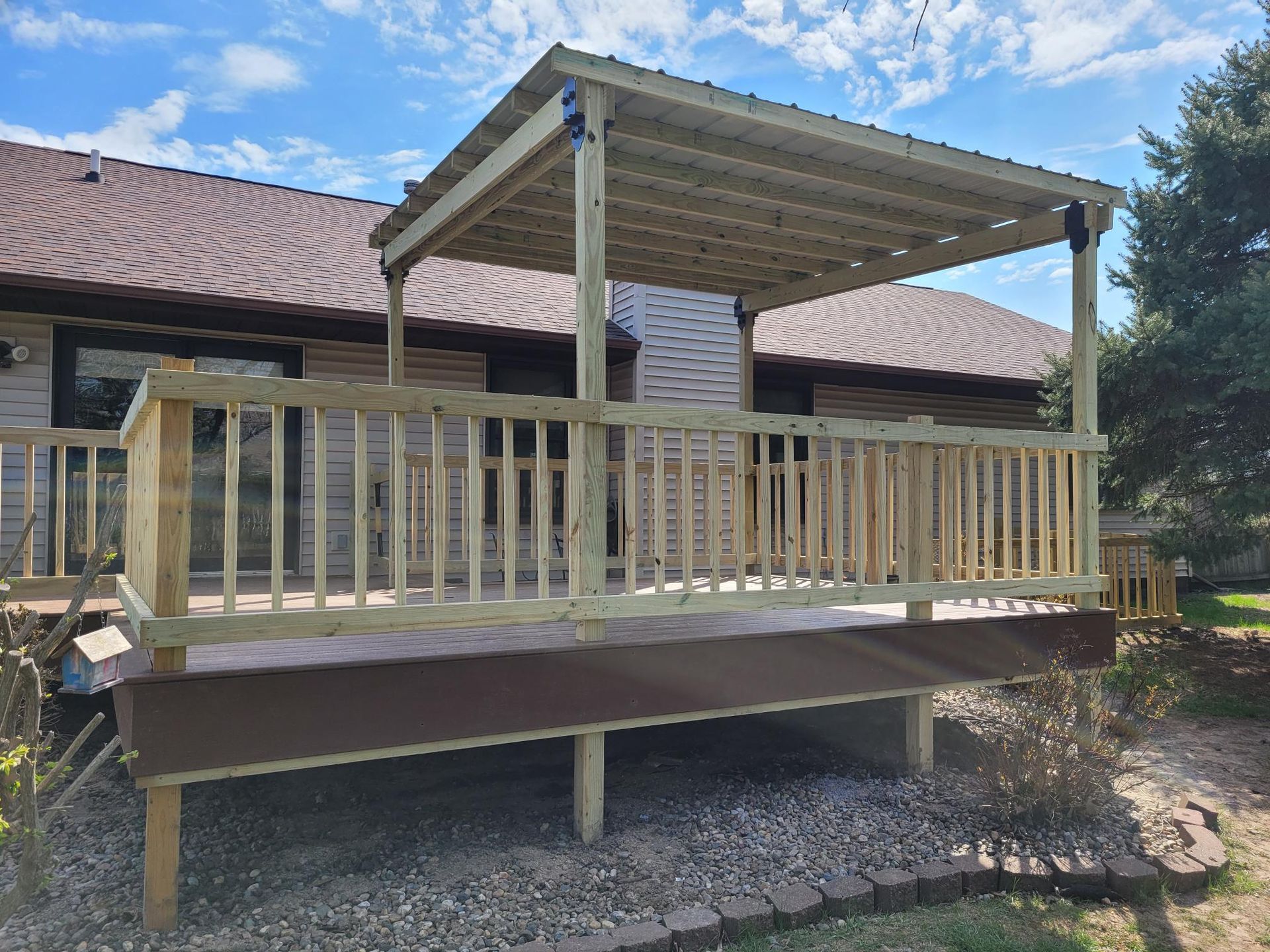 Wooden deck with railing and a shaded pergola attached to a house, beside a gravel yard.