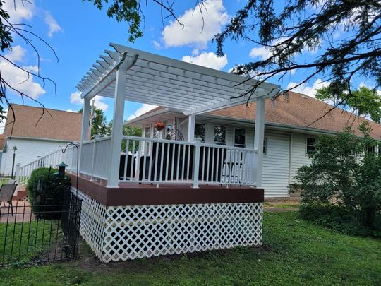 White porch with lattice skirting on a house, under a pergola, in a grassy yard under blue sky