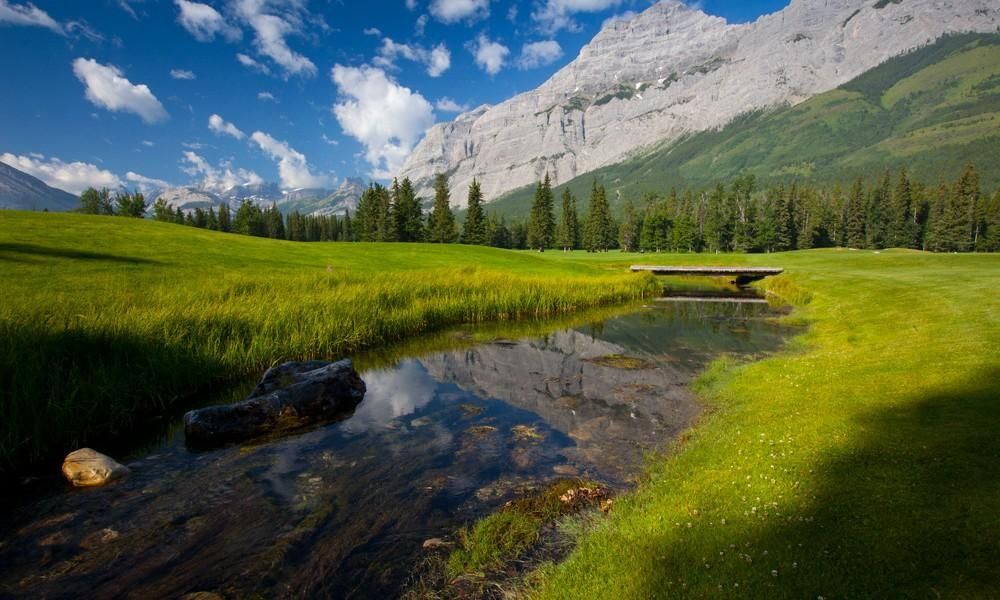 A bridge over a stream in a grassy field with mountains in the background.