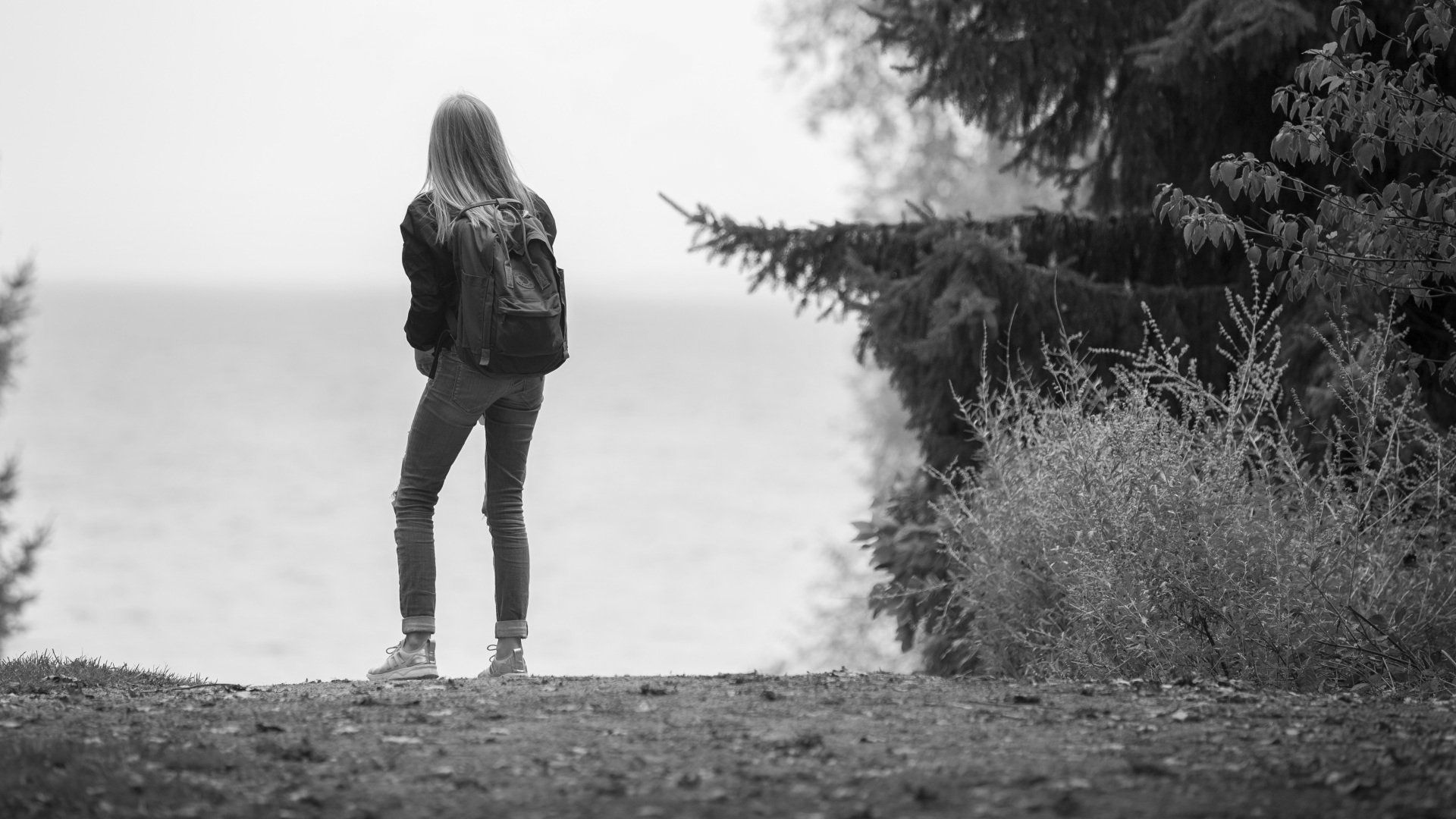 A woman with a backpack is standing on a hill overlooking the ocean.