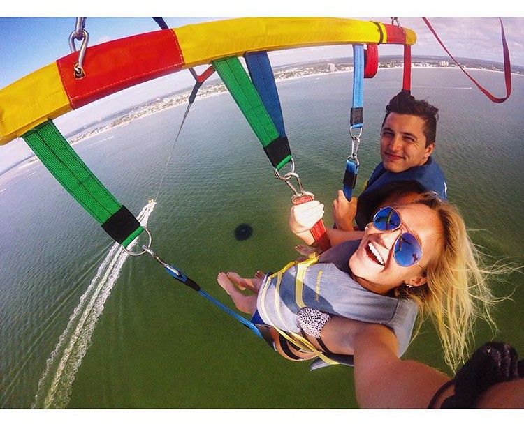 Two people hang gliding over green water, smiling in a colorful tandem harness.