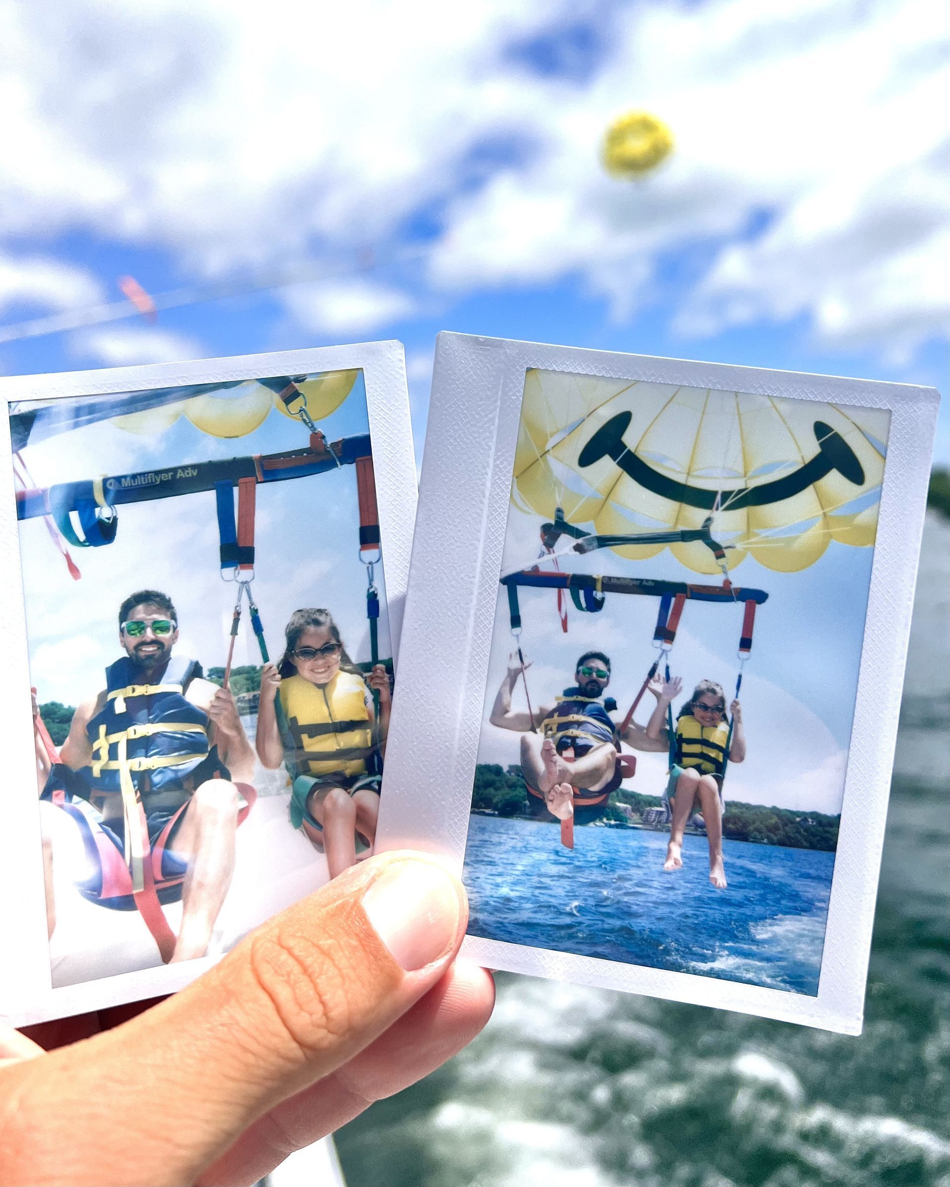 Hand holding two small photos of people parasailing over the ocean under a blue sky