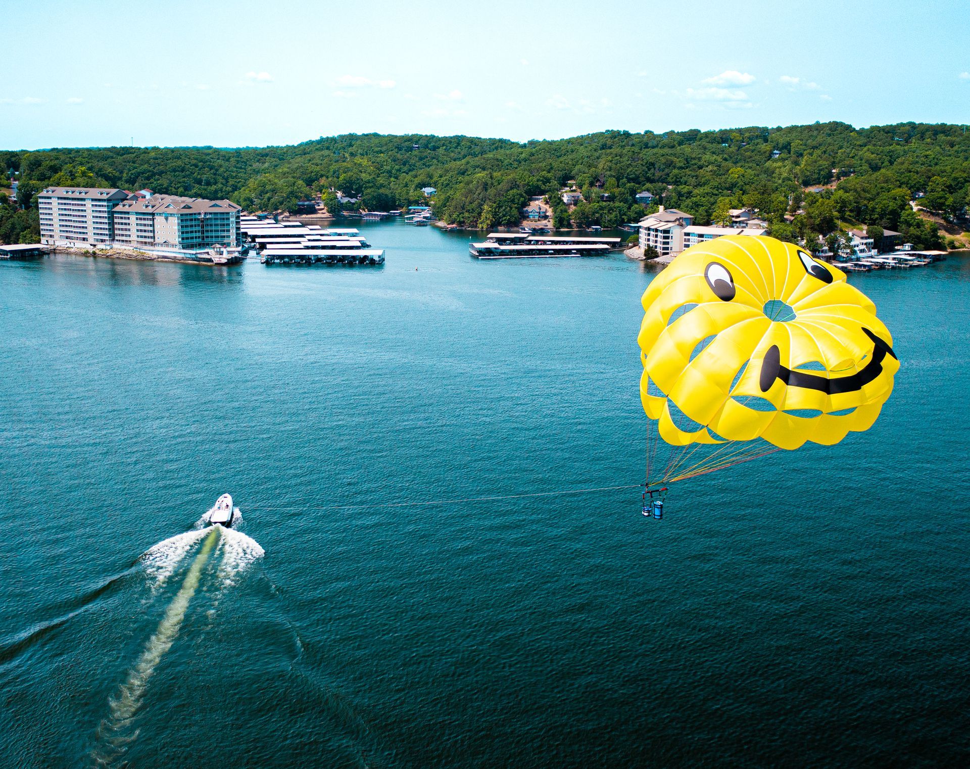 Parasailer over blue water near a wooded shoreline, with boats and buildings in the distance.
