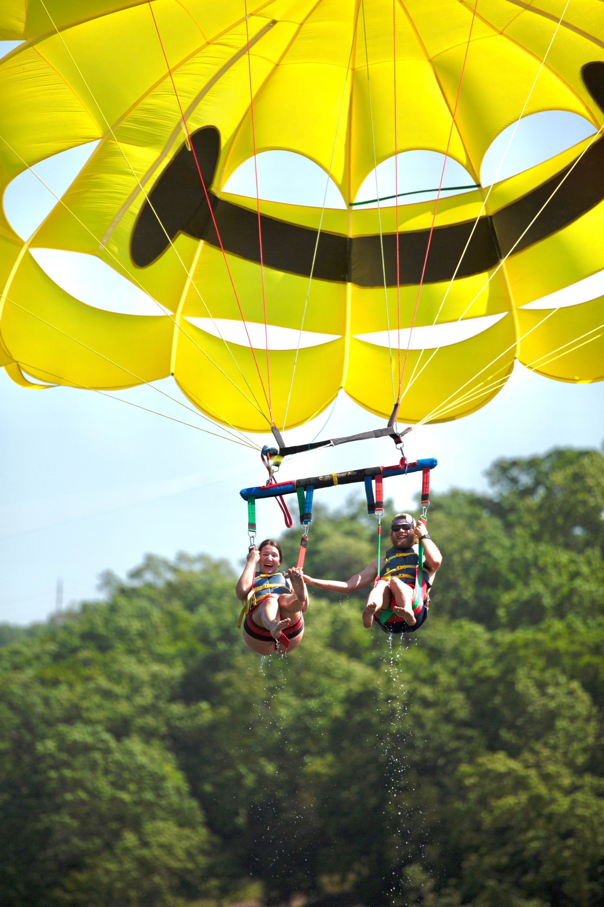 Two people parasailing under a bright yellow canopy above a forested shoreline