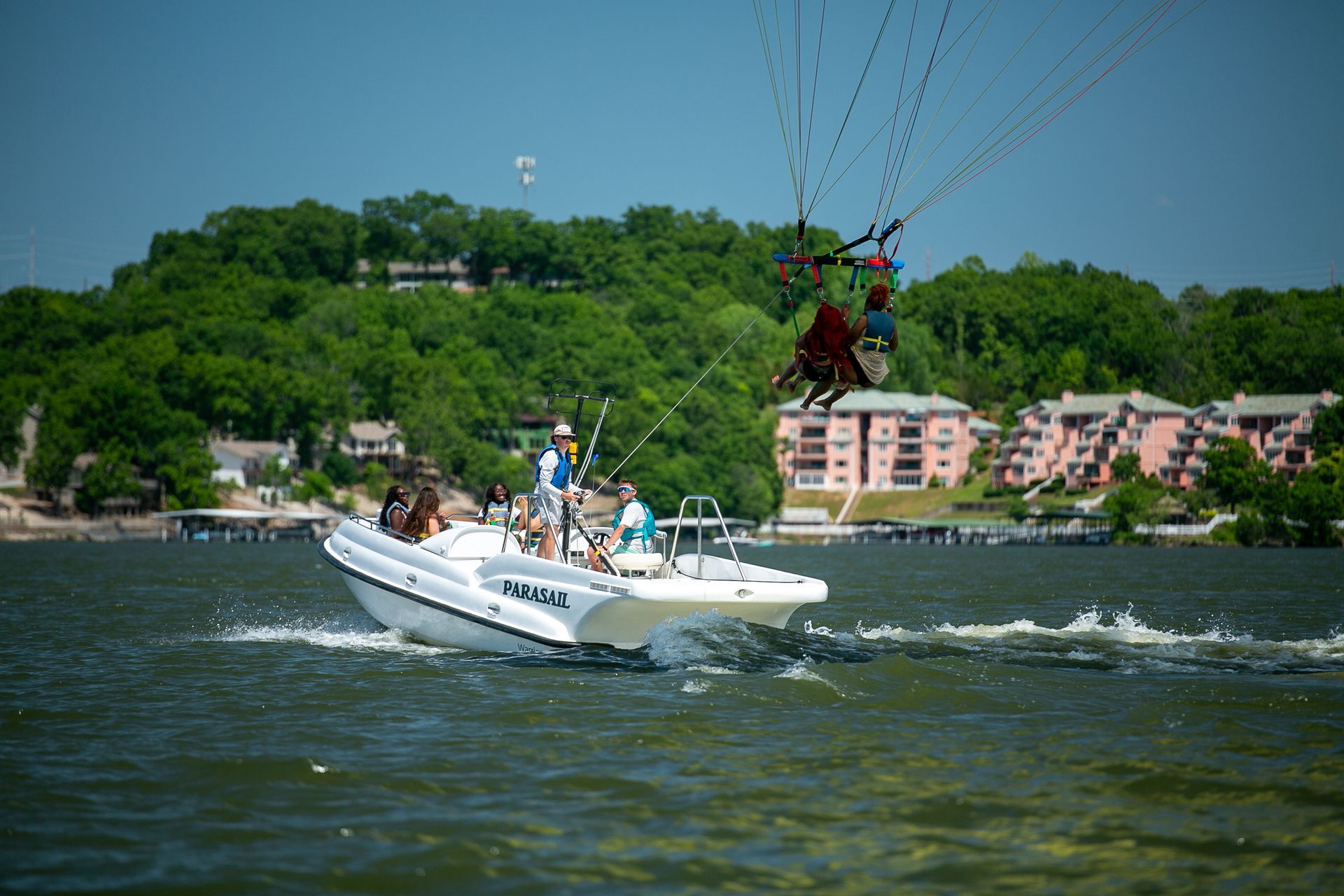Speedboat towing a parasail over choppy water with a green hillside and lakeside buildings in the background