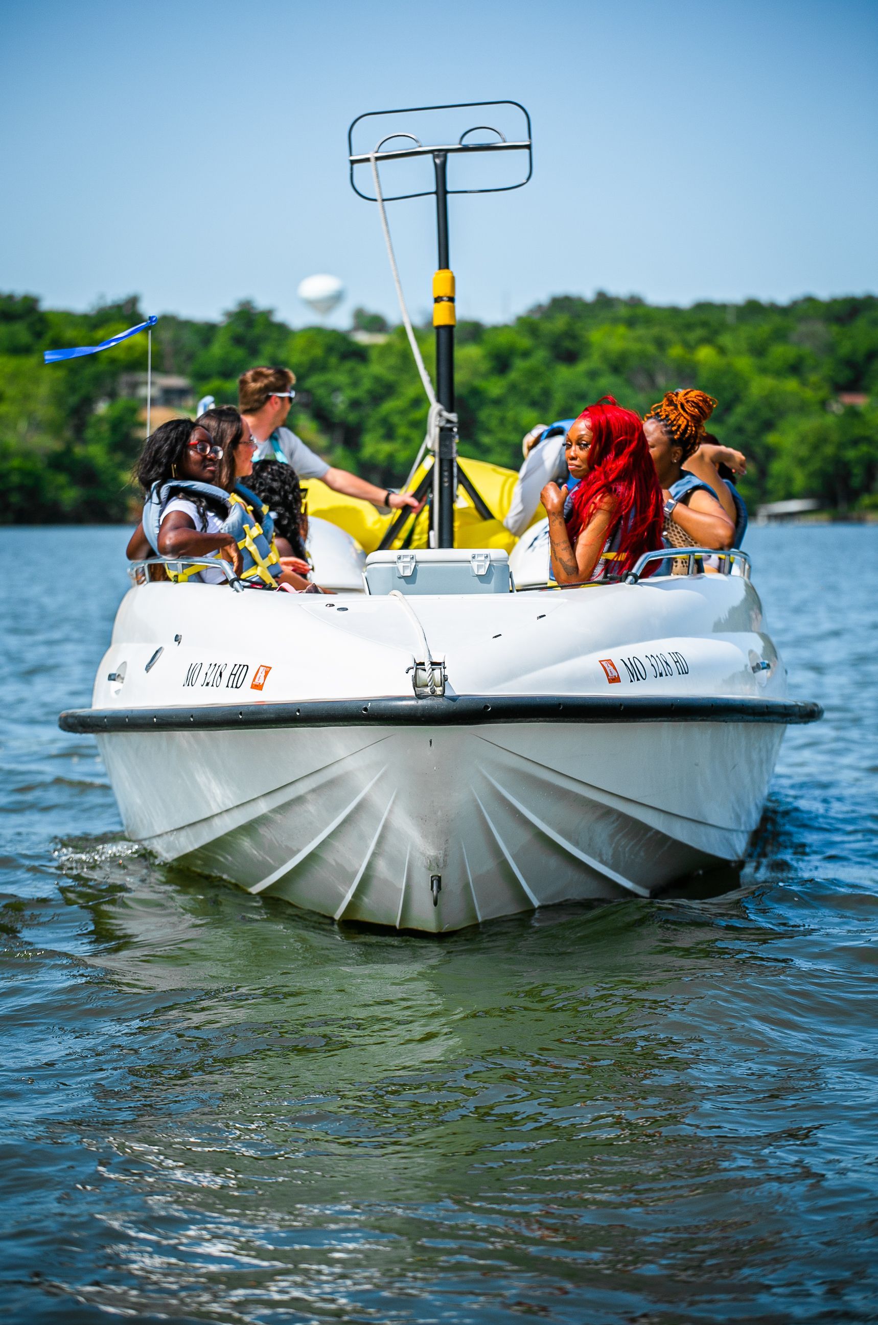Small white boat with people on a lake, seen from the front under a sunny blue sky.