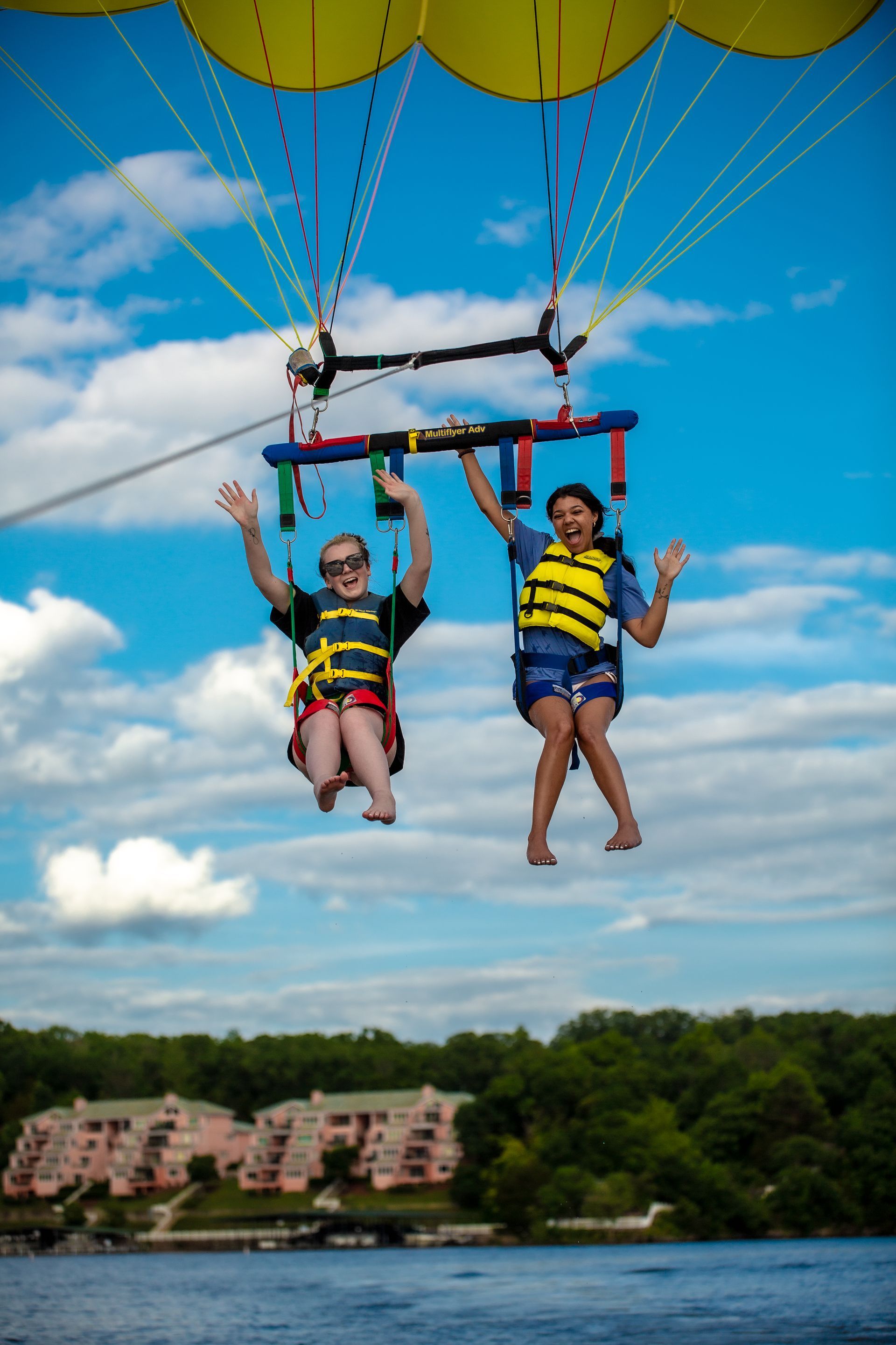 Two people parasailing above a lake under yellow canopies, with buildings and trees in the background.