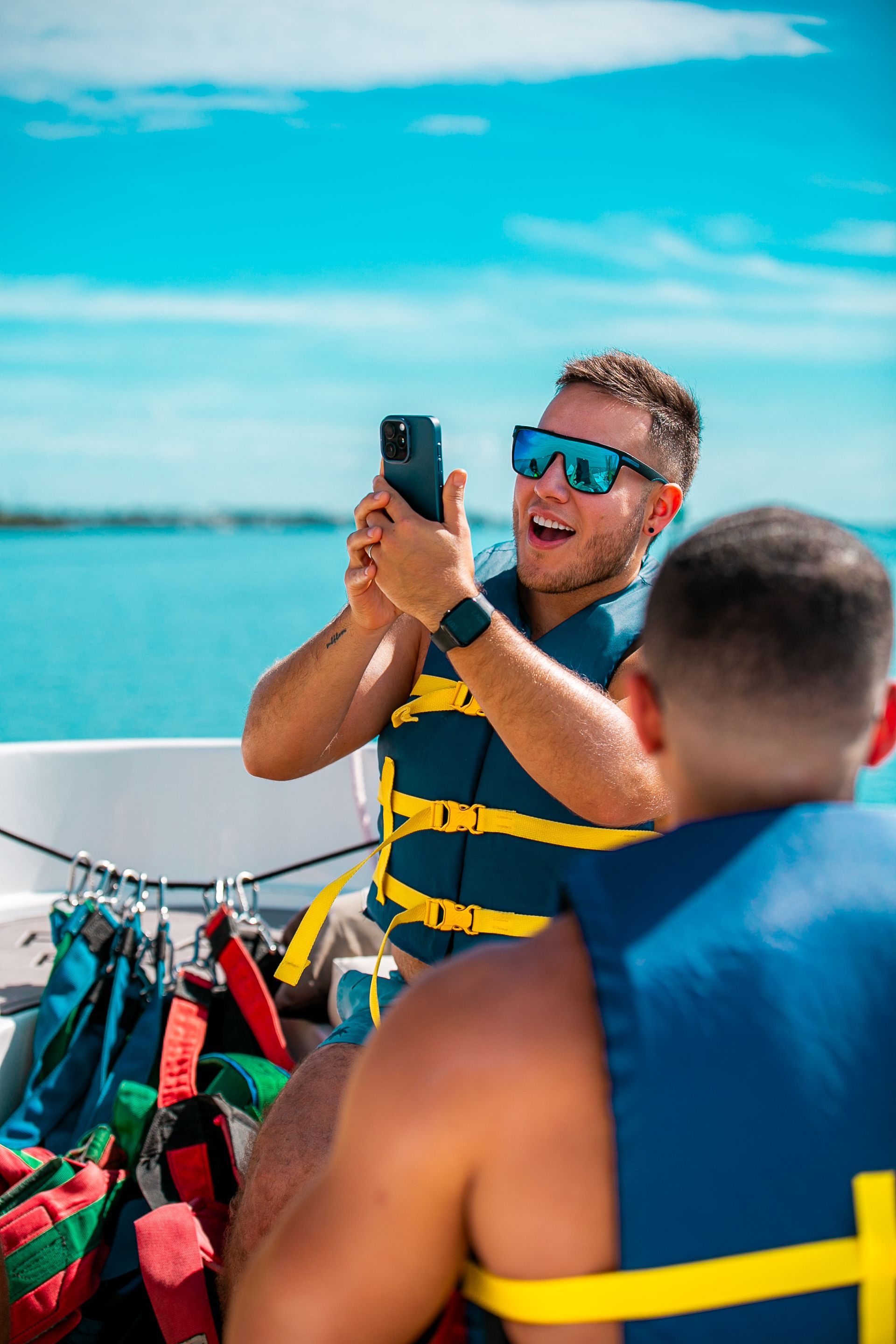 Man in blue shirt and sunglasses taking a photo on a boat, with turquoise water and a bright sky behind him