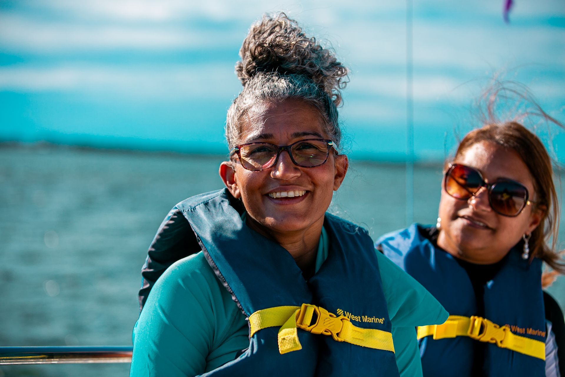 Two smiling people in teal life vests on a boat, with water in the background.