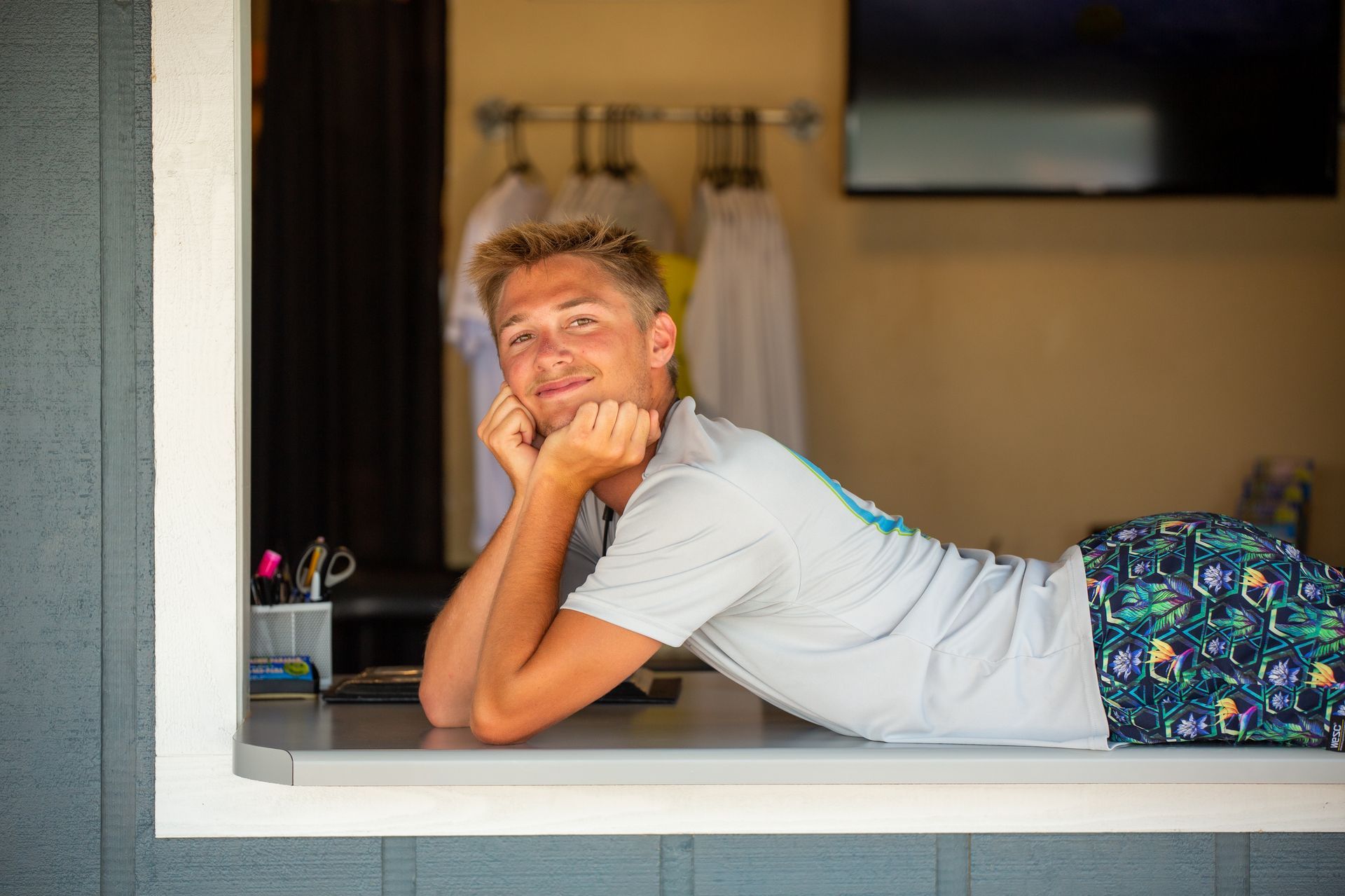 Person leaning on a windowsill, smiling in a white shirt with colorful shorts.