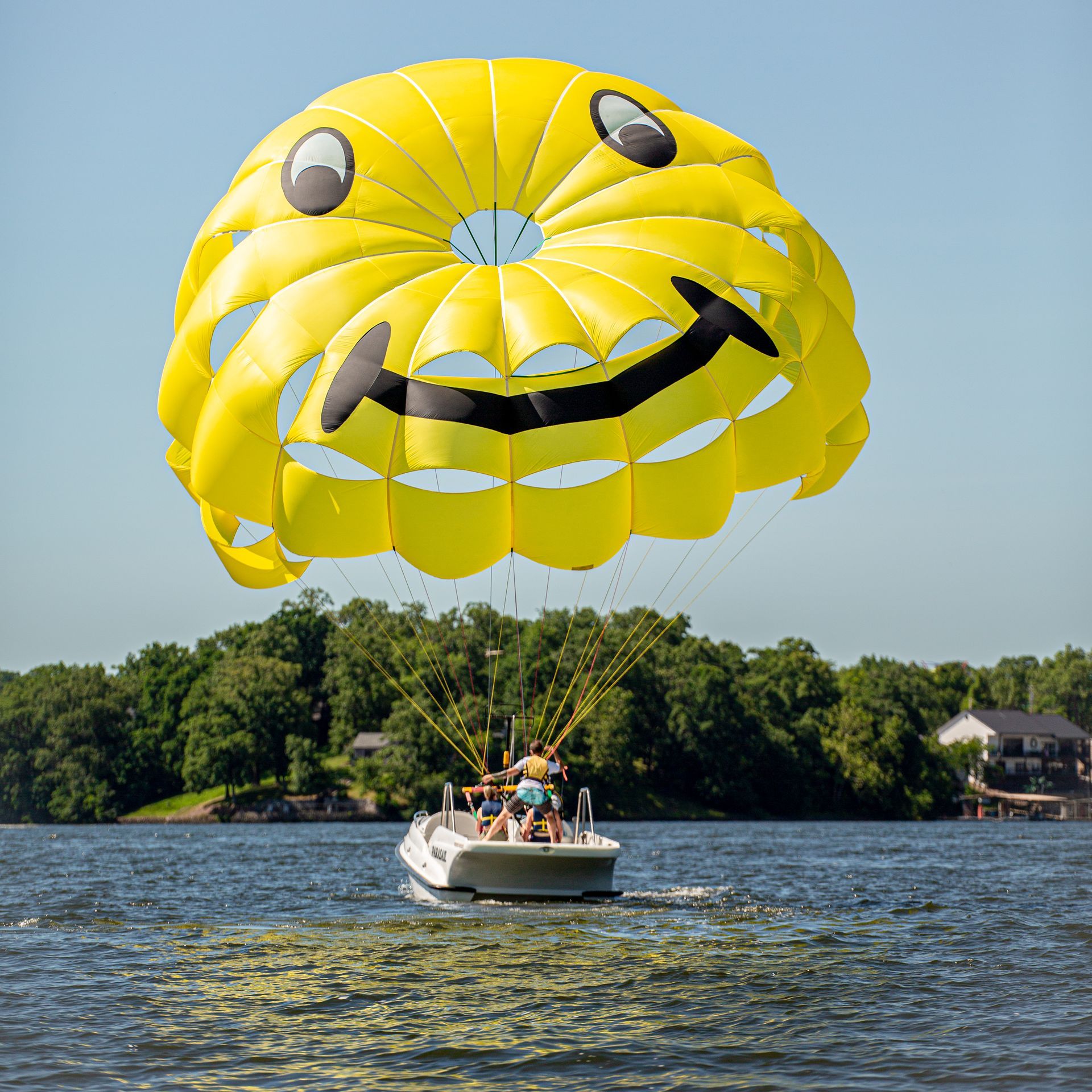 Boat parasailing with a large yellow smiley parachute over a lake near a wooded shore