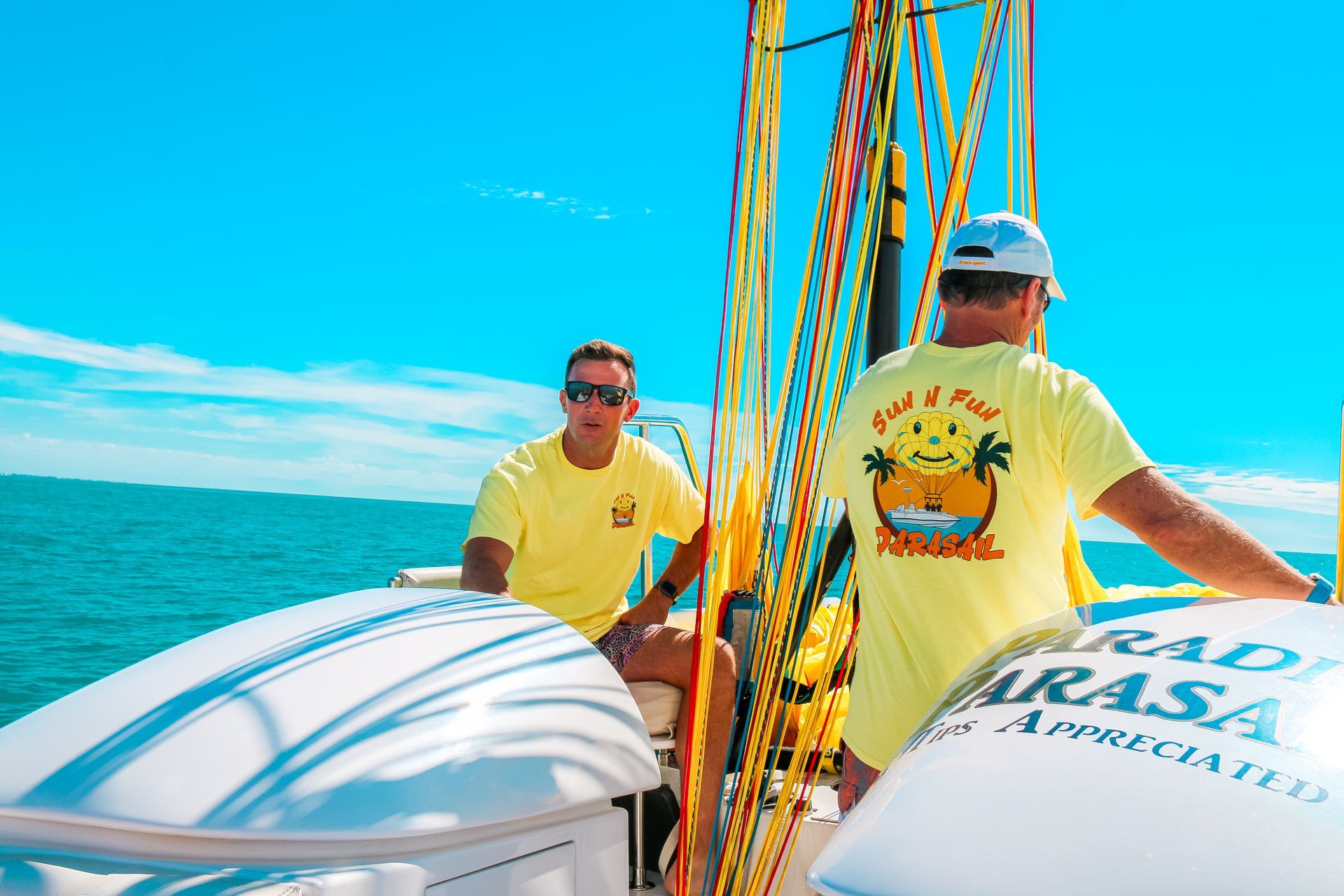 Two people in yellow shirts on a sailboat deck, with ocean and blue sky in the background