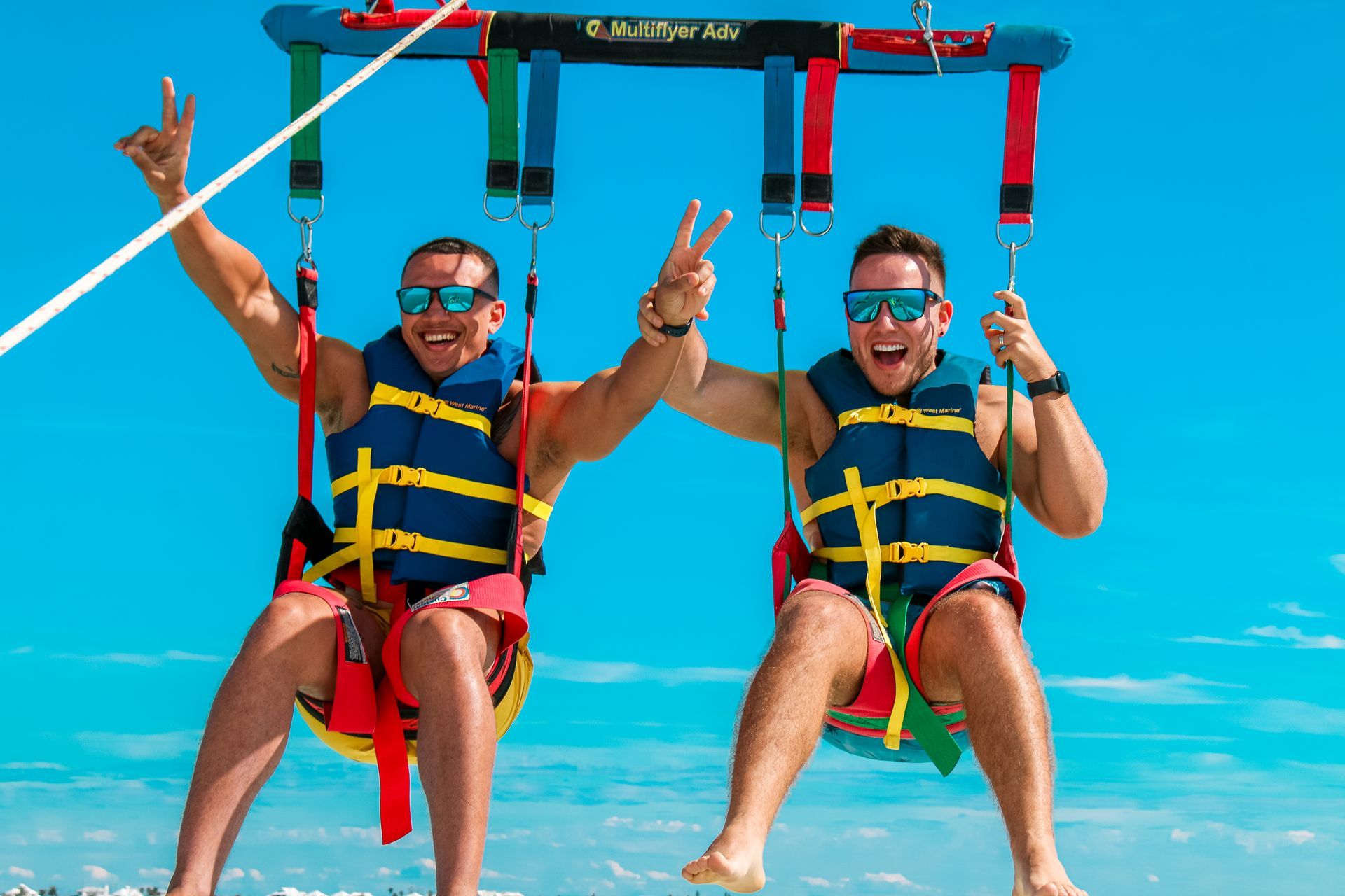 Two people parasailing over bright blue water, smiling and raising their arms.