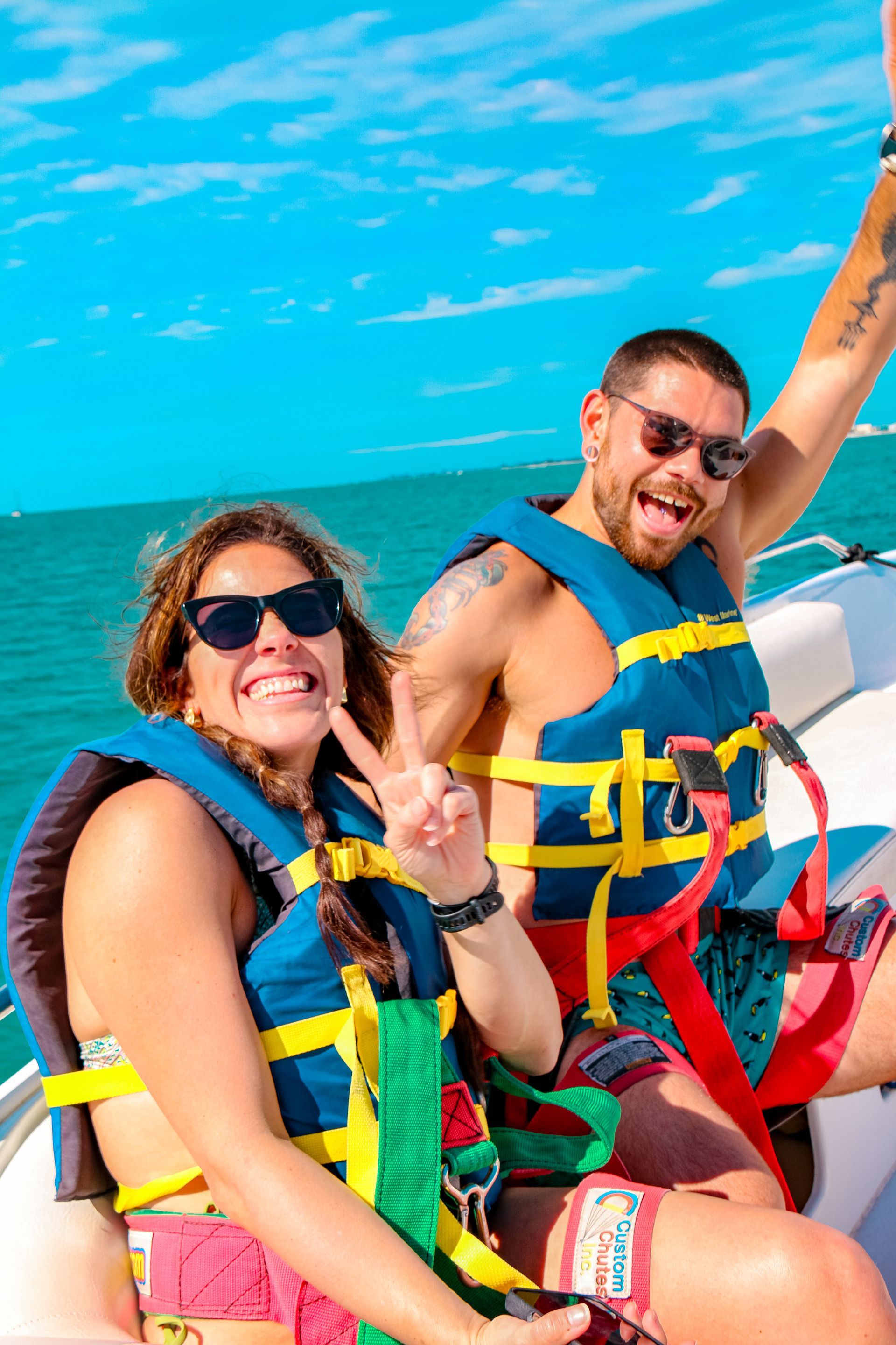 Two people smiling in life vests and harnesses on a boat, with ocean and blue sky behind them