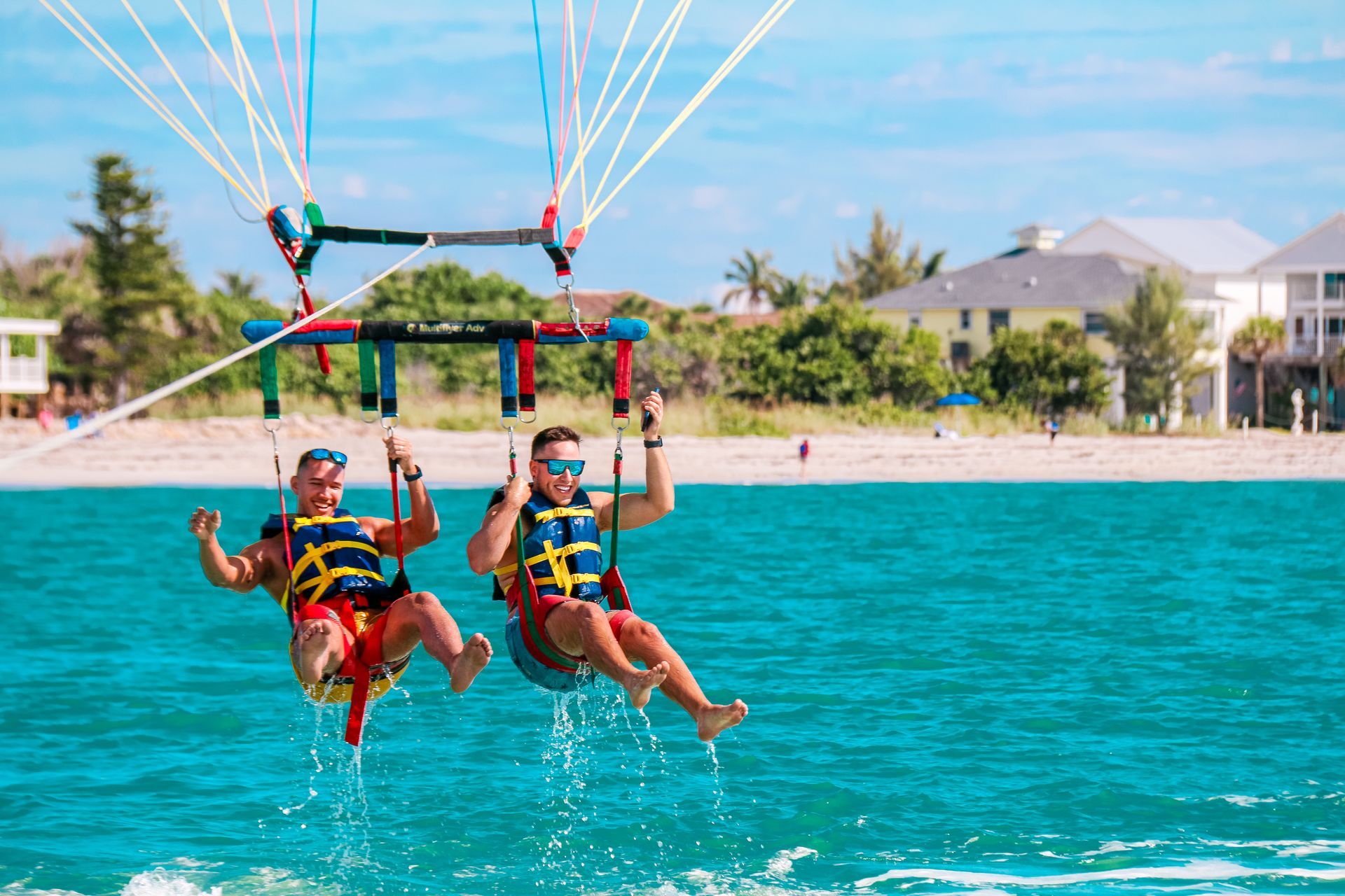 Two people parasailing over turquoise water near a tropical shoreline.