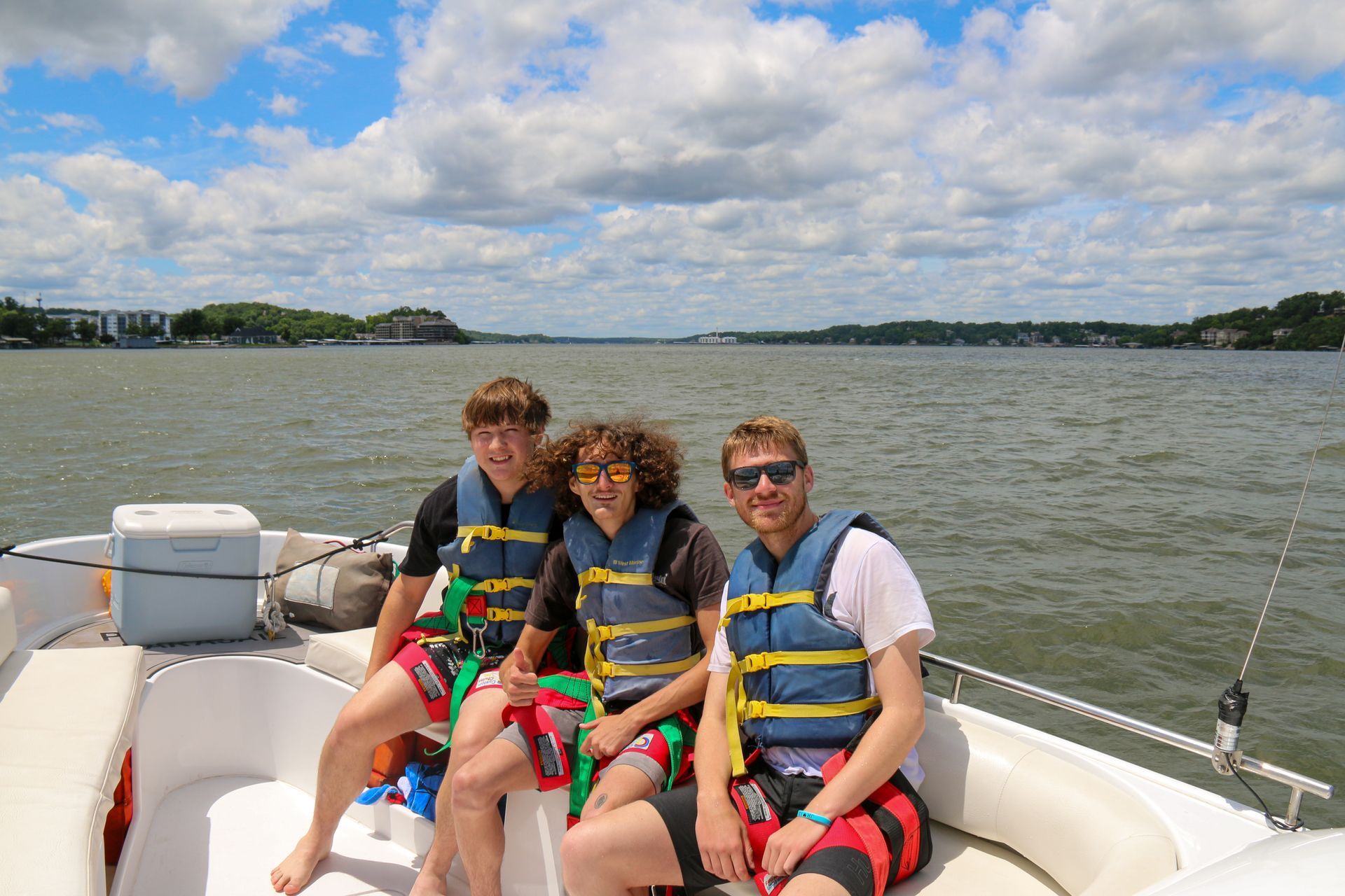 Three people in life jackets sit on a boat on a lake under a cloudy sky.