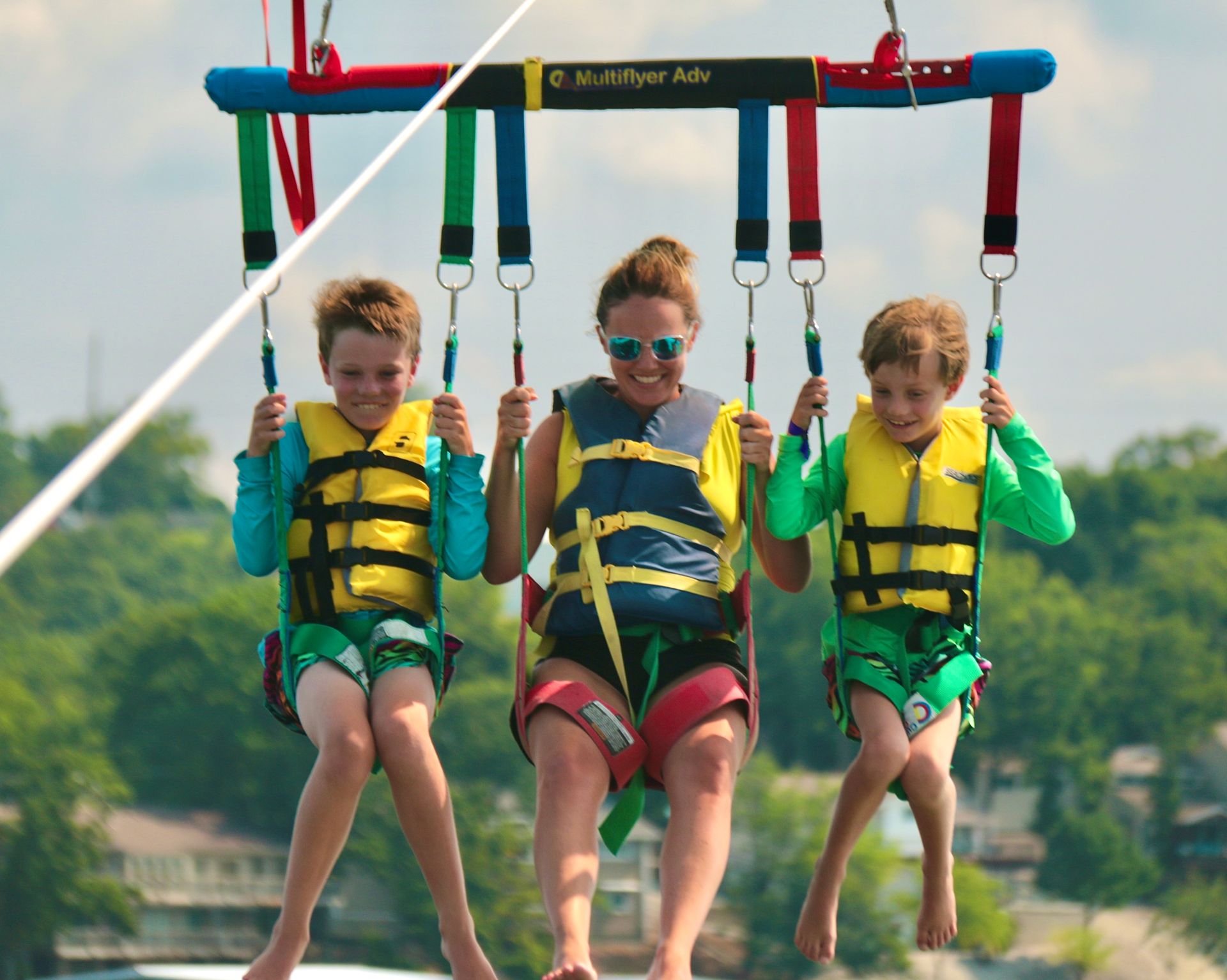 Three children in life jackets zip lining together over water, smiling and holding the harnesses.