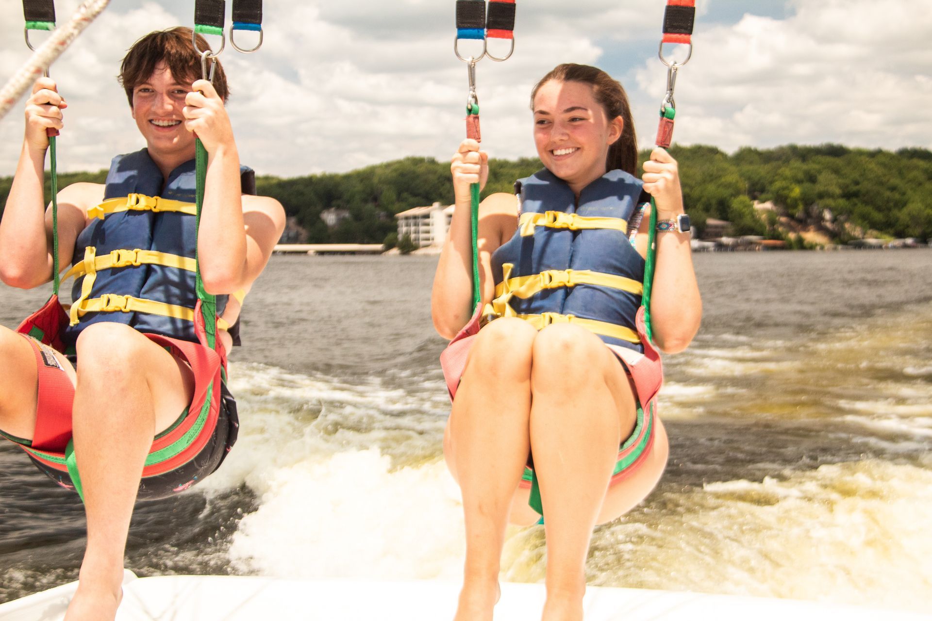 Two people in life jackets riding a water swing over a lake, smiling and splashing through the air