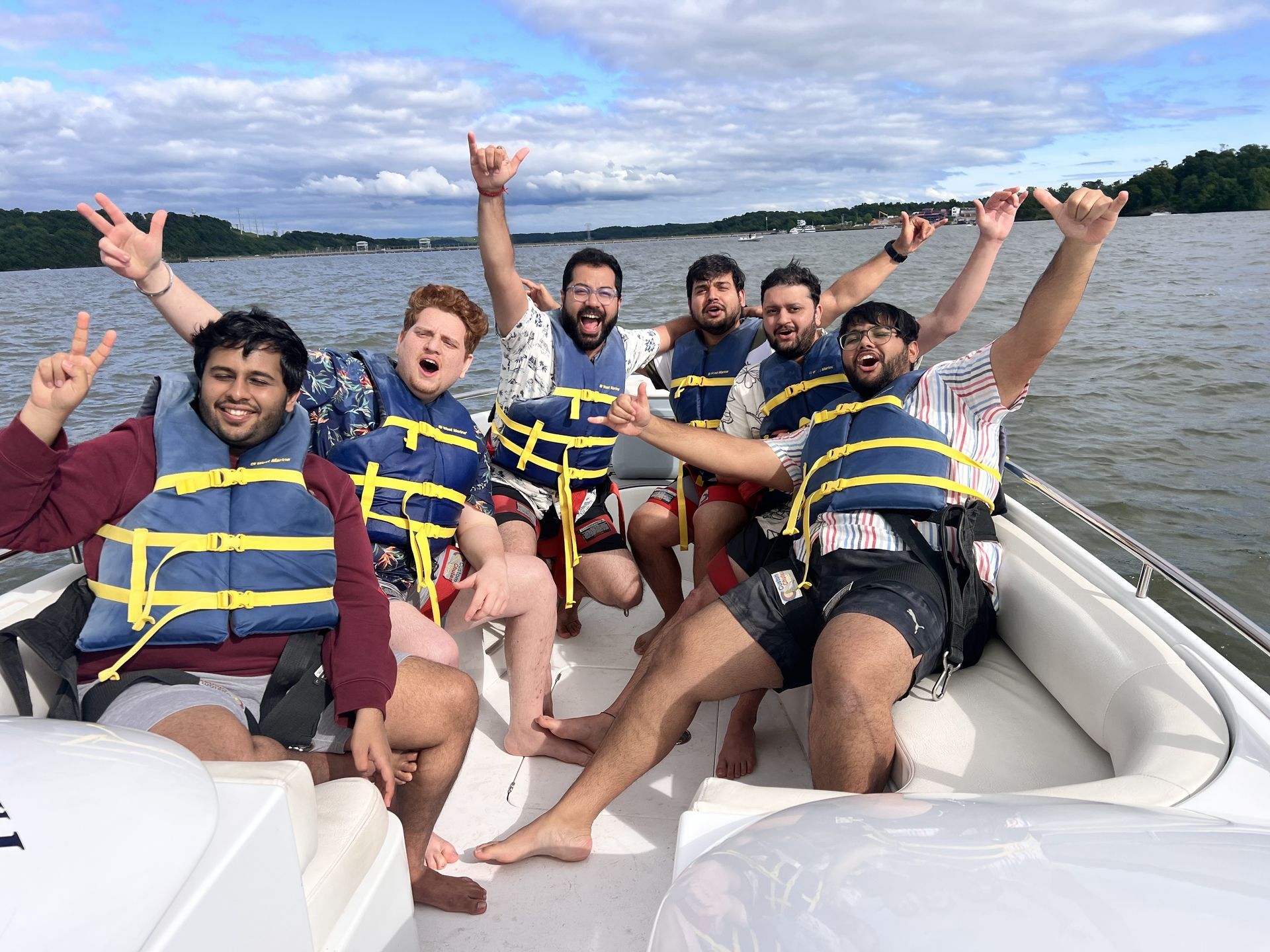 Six friends in life jackets cheer on a speedboat on a lake under a blue sky.