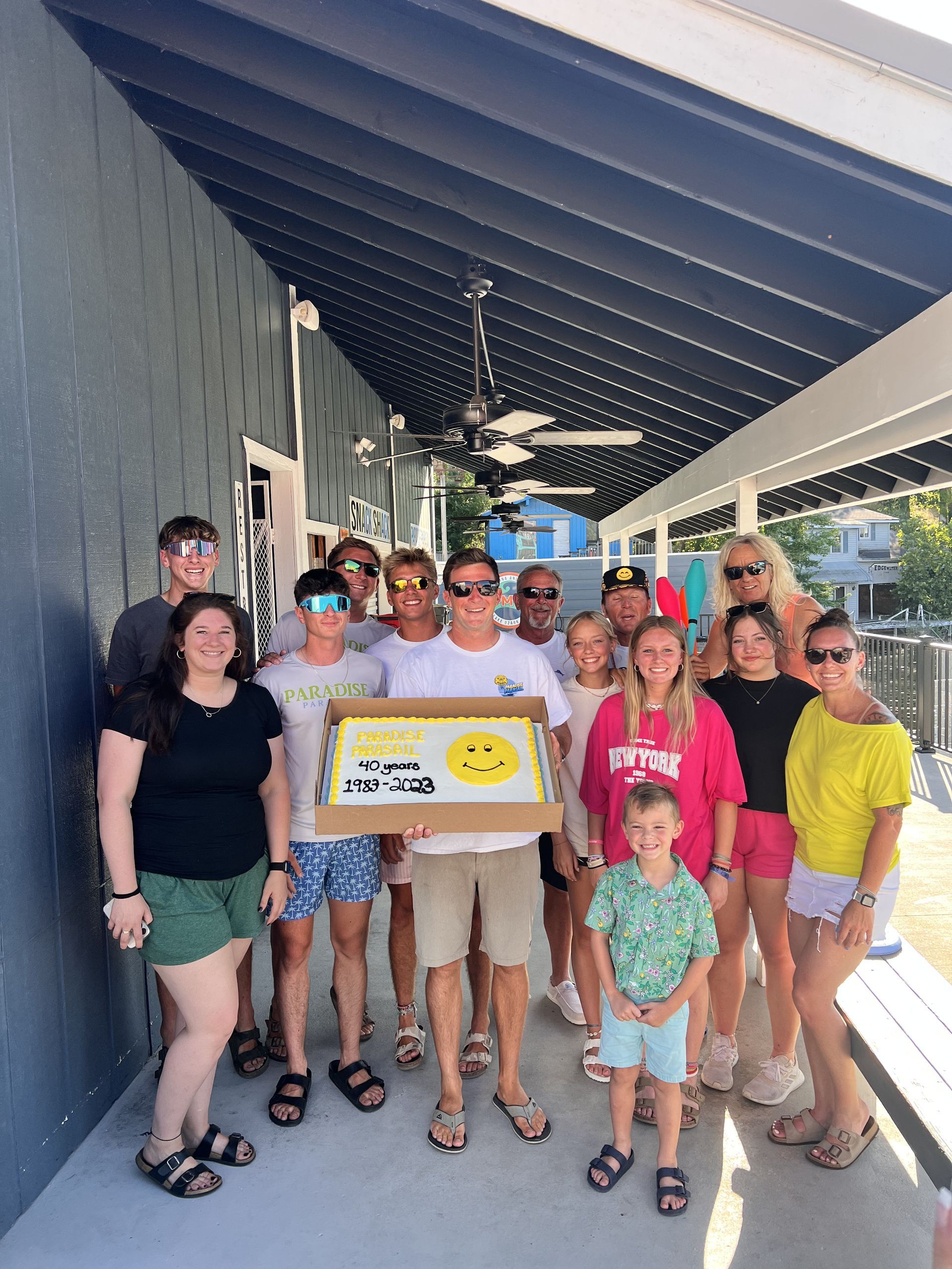 Group posing on a shaded walkway, holding a framed certificate and yellow ribbon banner.