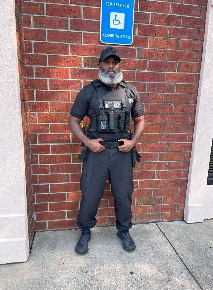Man in security uniform stands in front of a brick wall, under a handicap parking sign.