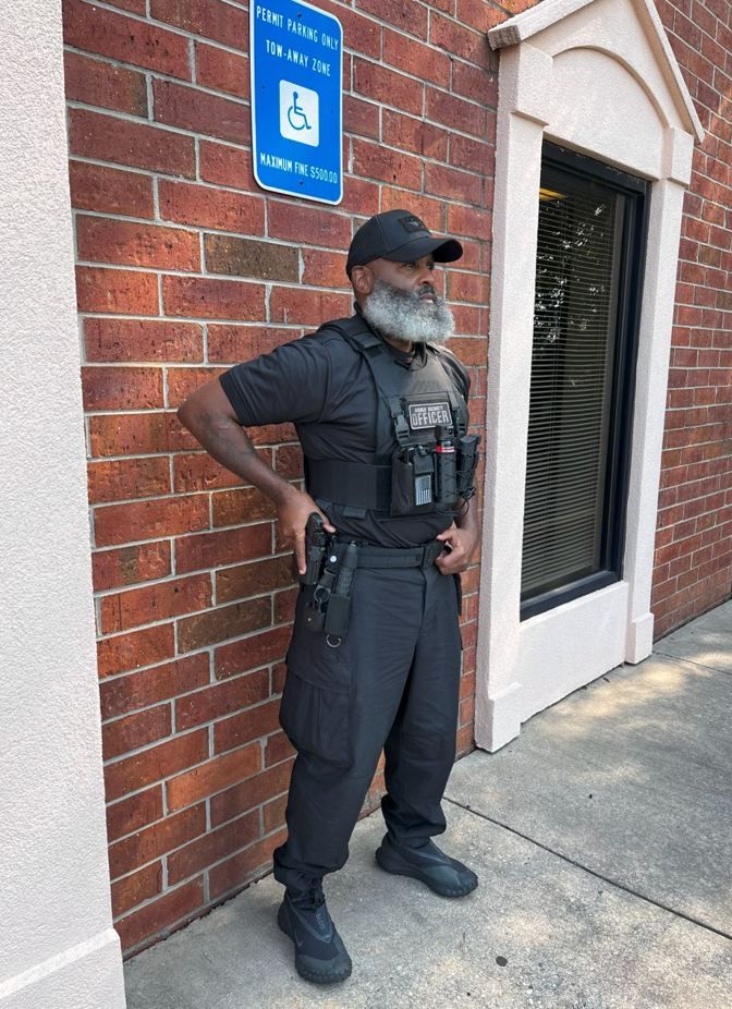 Man in tactical gear stands near a brick building. He has a beard, is wearing a hat, and a holster.