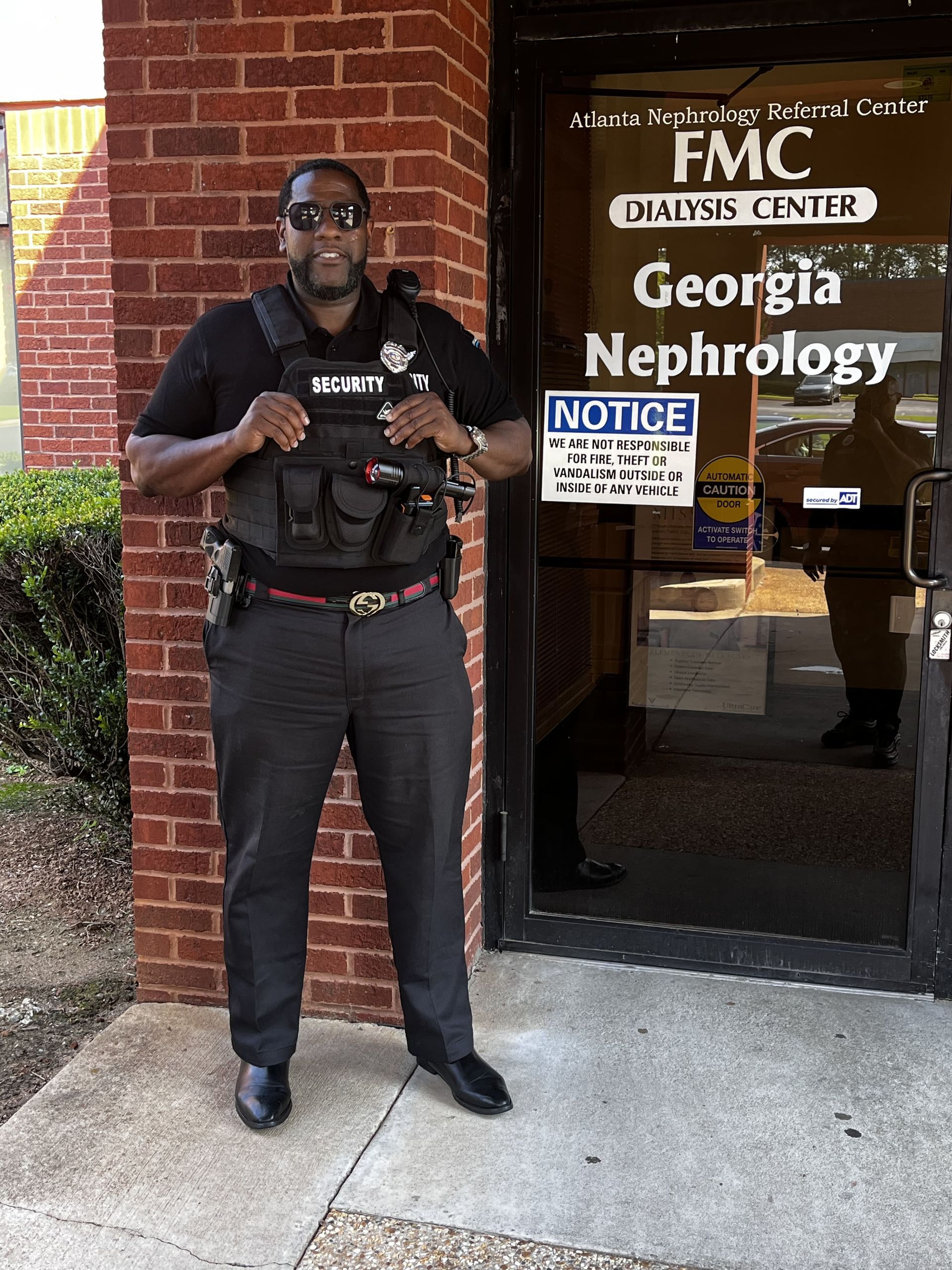 Security guard stands at the entrance of a dialysis center. He wears a vest, sunglasses, and a stern expression.