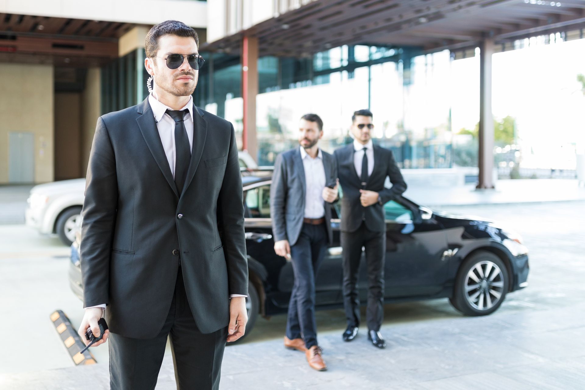 Bodyguards in black suits and sunglasses escorting men near a black car, outdoors.