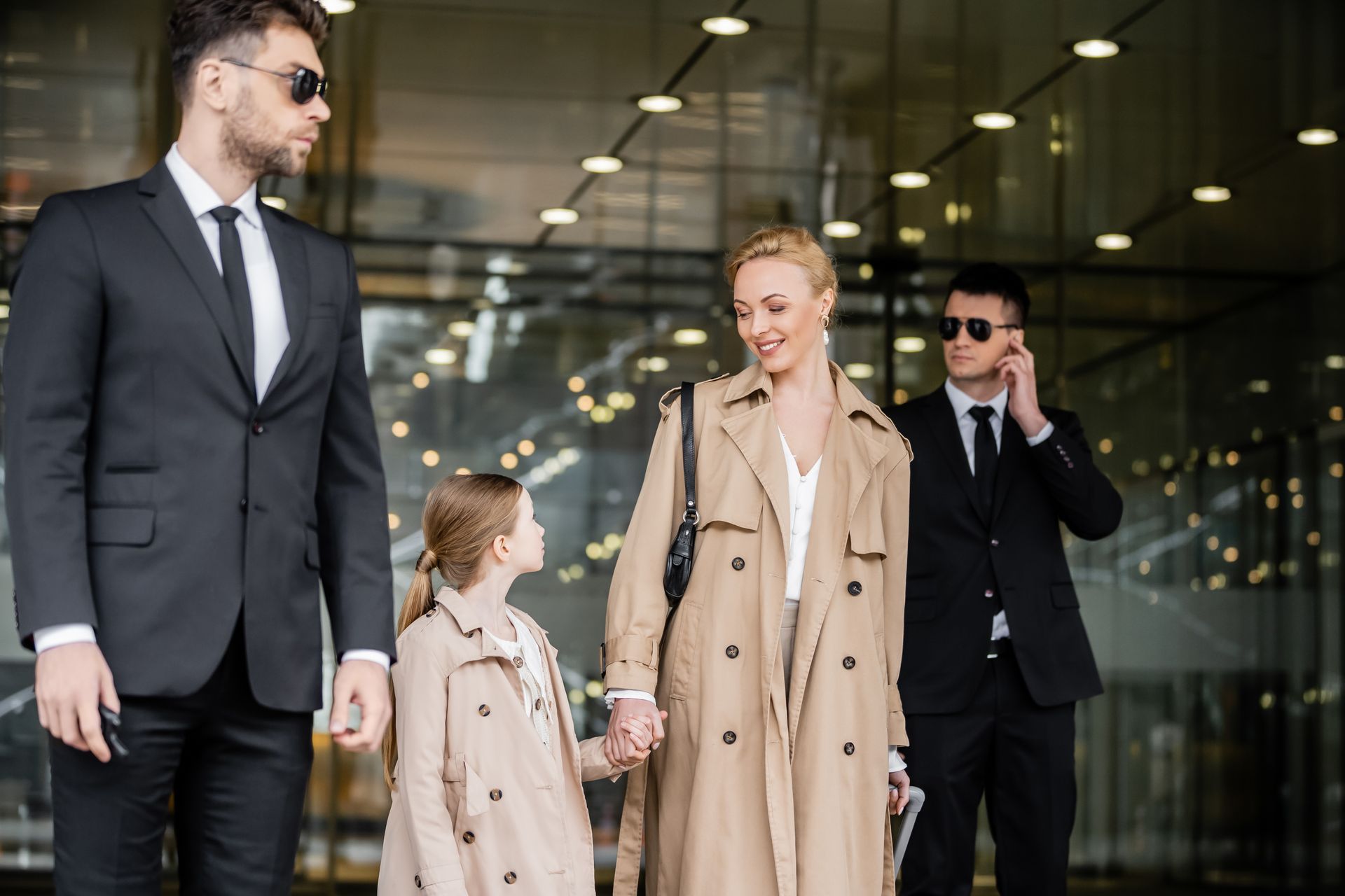 A woman and child, hand-in-hand, exit a building followed by two bodyguards, all wearing sunglasses.