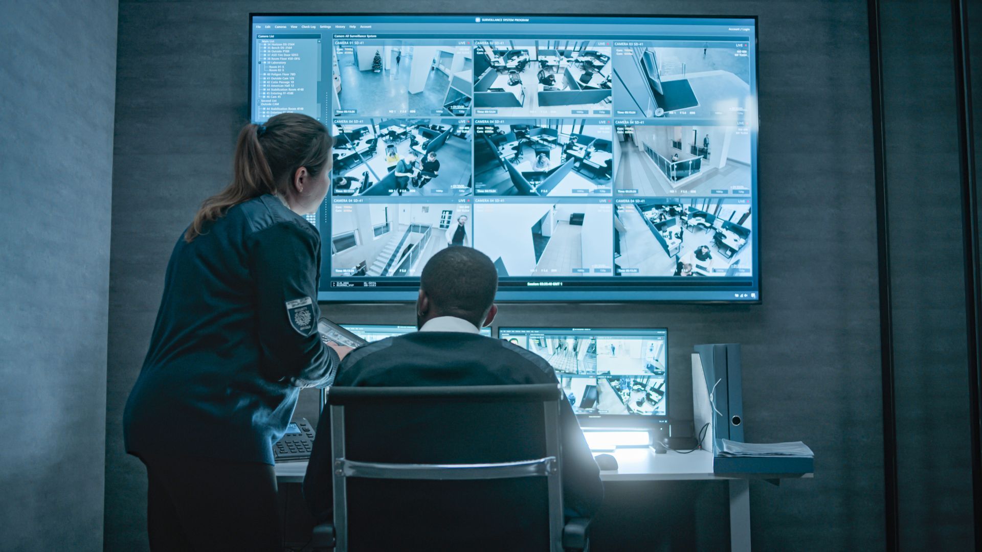 Two people in a security room watching surveillance monitors. A man sits at a desk, a woman stands nearby.