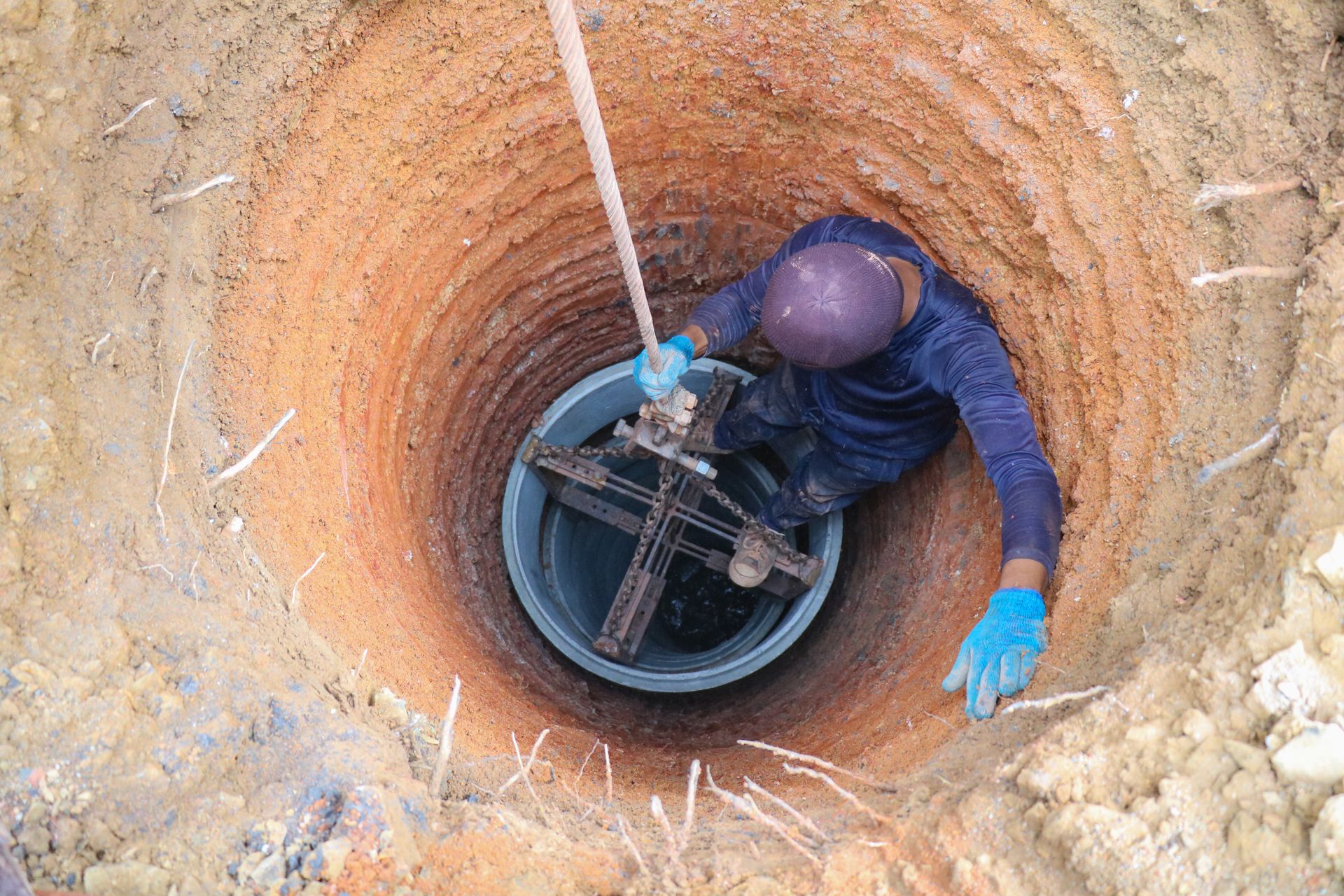 Worker in a deep utility hole handling equipment on a pipe, wearing a hard hat and blue gloves.
