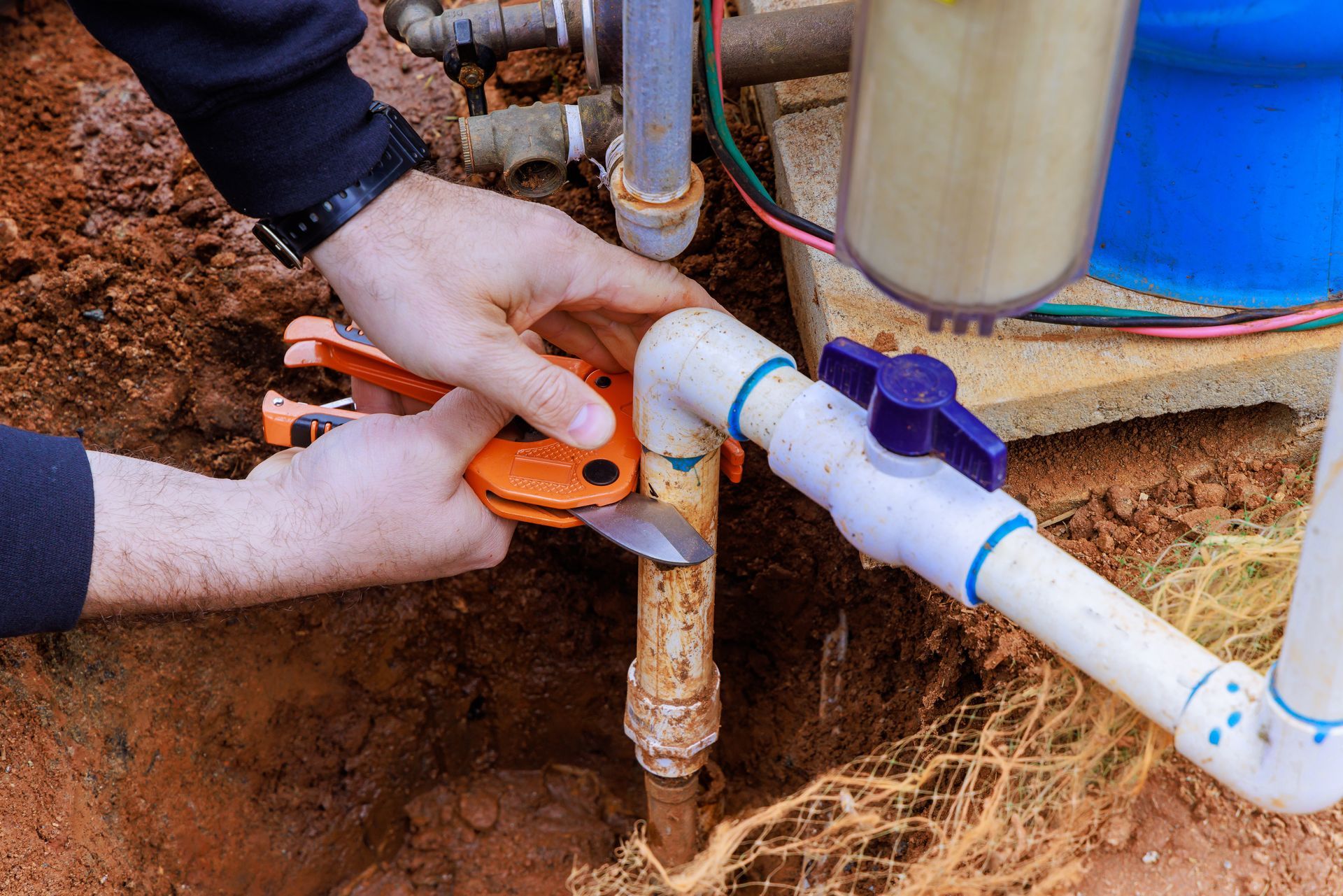 Hands using orange and silver pipe cutters on a PVC pipe outdoors near a water system