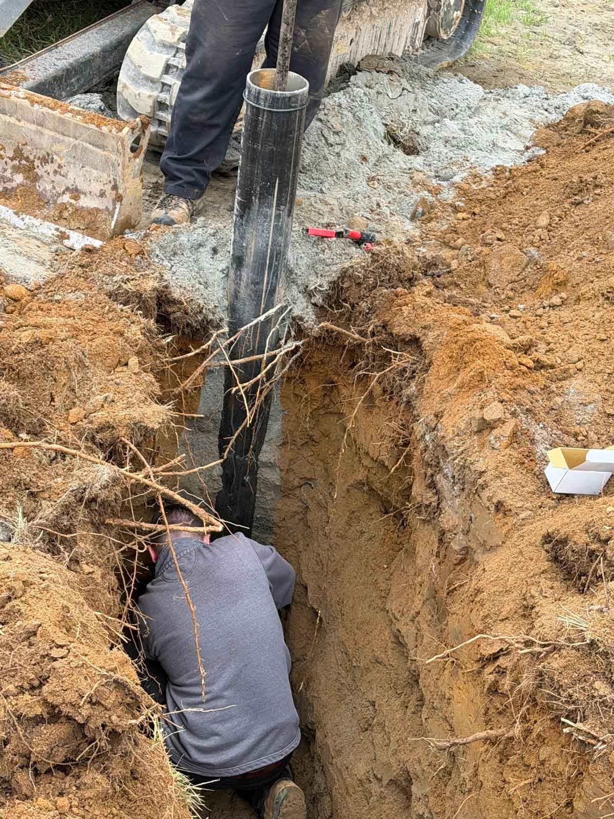 Worker in a deep trench beside a black pole, handling roots and soil during installation work.
