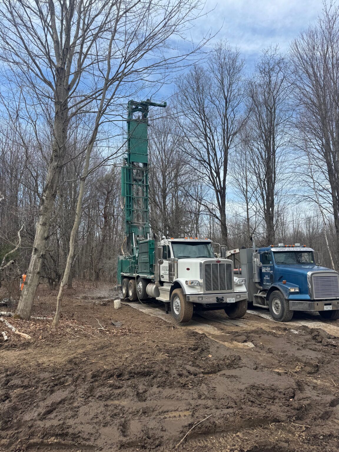 Two trucks beside a tall green drilling rig in a muddy wooded area