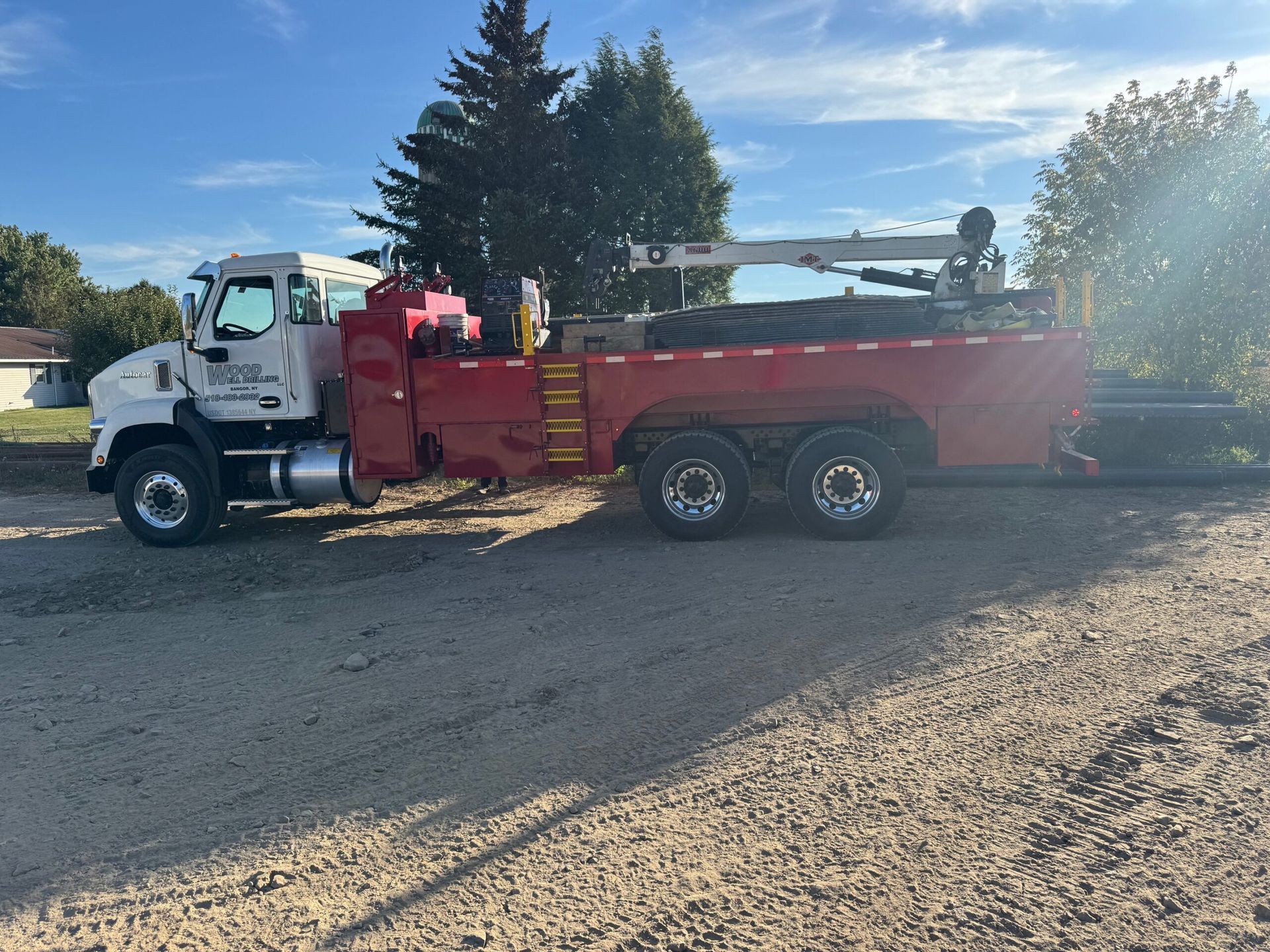 White truck with red dump bed parked on gravel in a sunny lot, with trees and a clear sky behind it