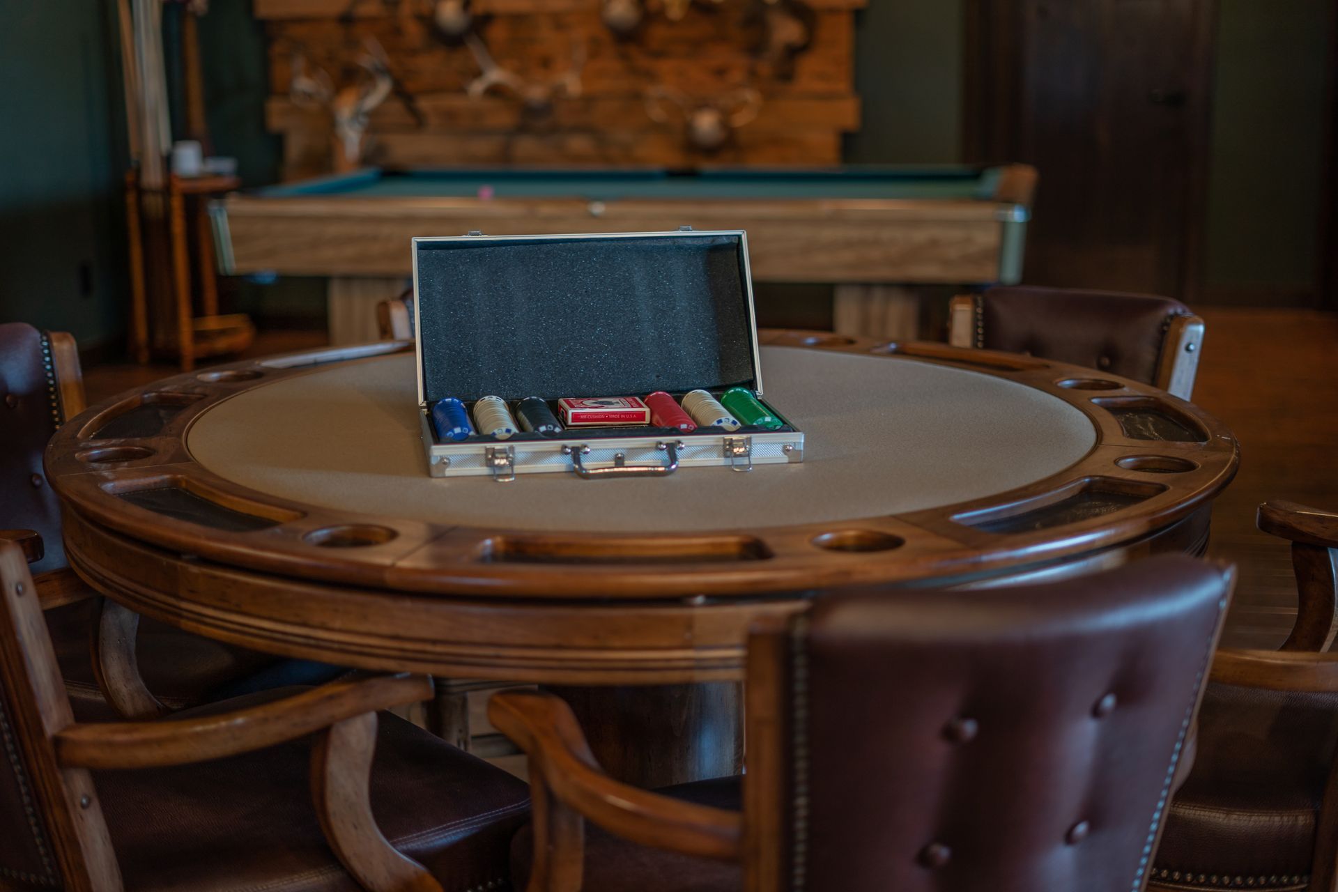 A poker table with a briefcase of poker chips on it in front of a pool table.