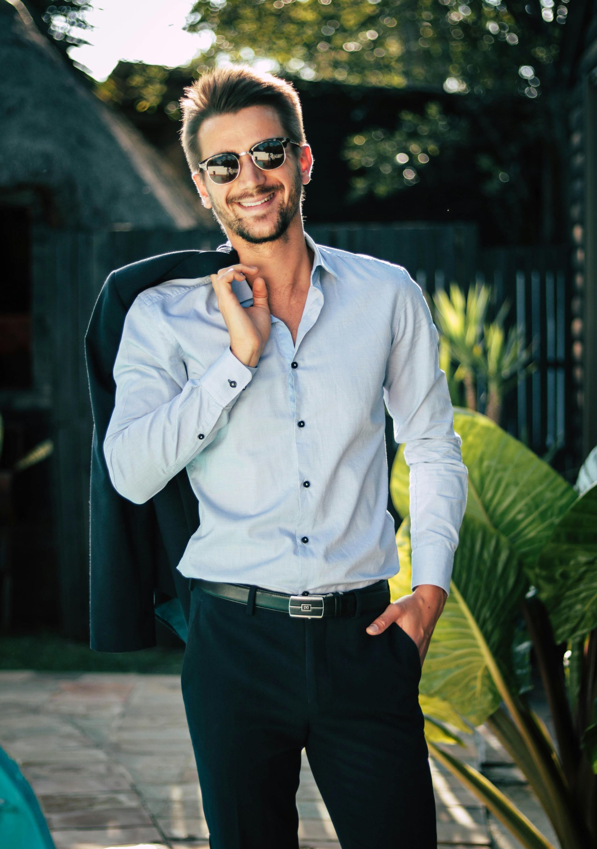 A man wearing sunglasses and a white shirt is holding his jacket over his shoulder.