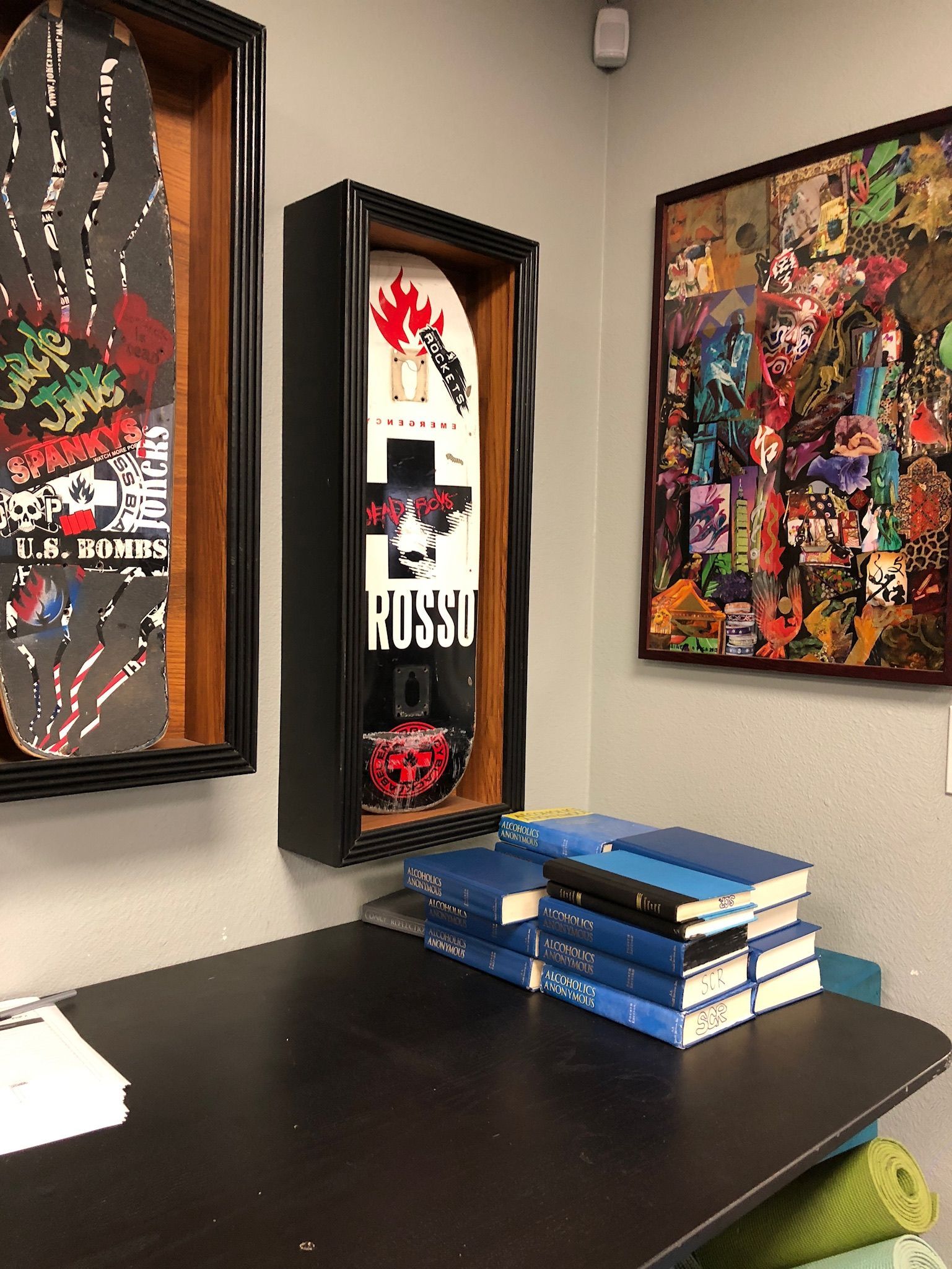 Skateboard art in framed boxes, on a gray wall above a desk with books.