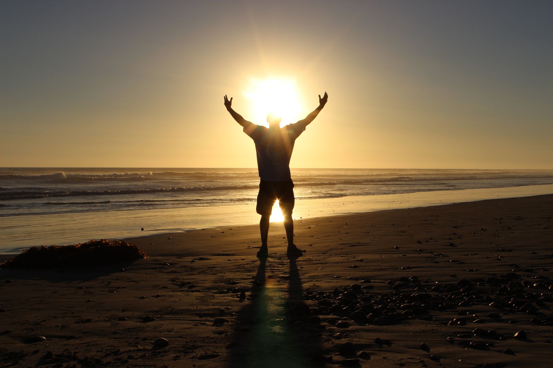 Man with arms raised on a beach at sunset, silhouetted by the sun's bright light.