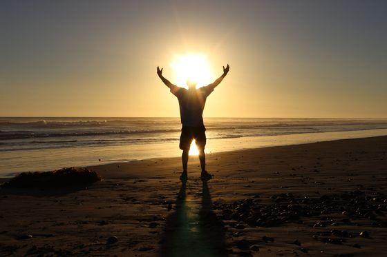 Man with arms raised on a beach at sunset, silhouetted by the sun's bright light.