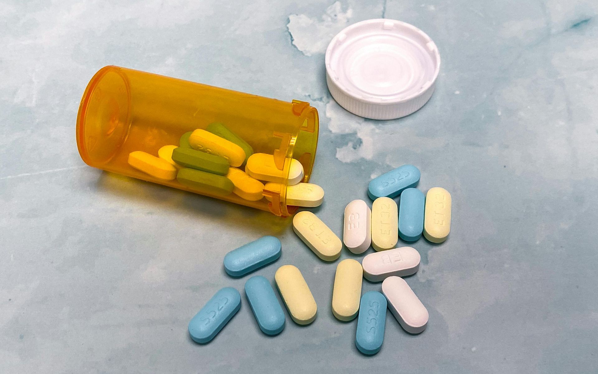 Pill bottle on its side, spilling various colored pills on a light blue surface.
