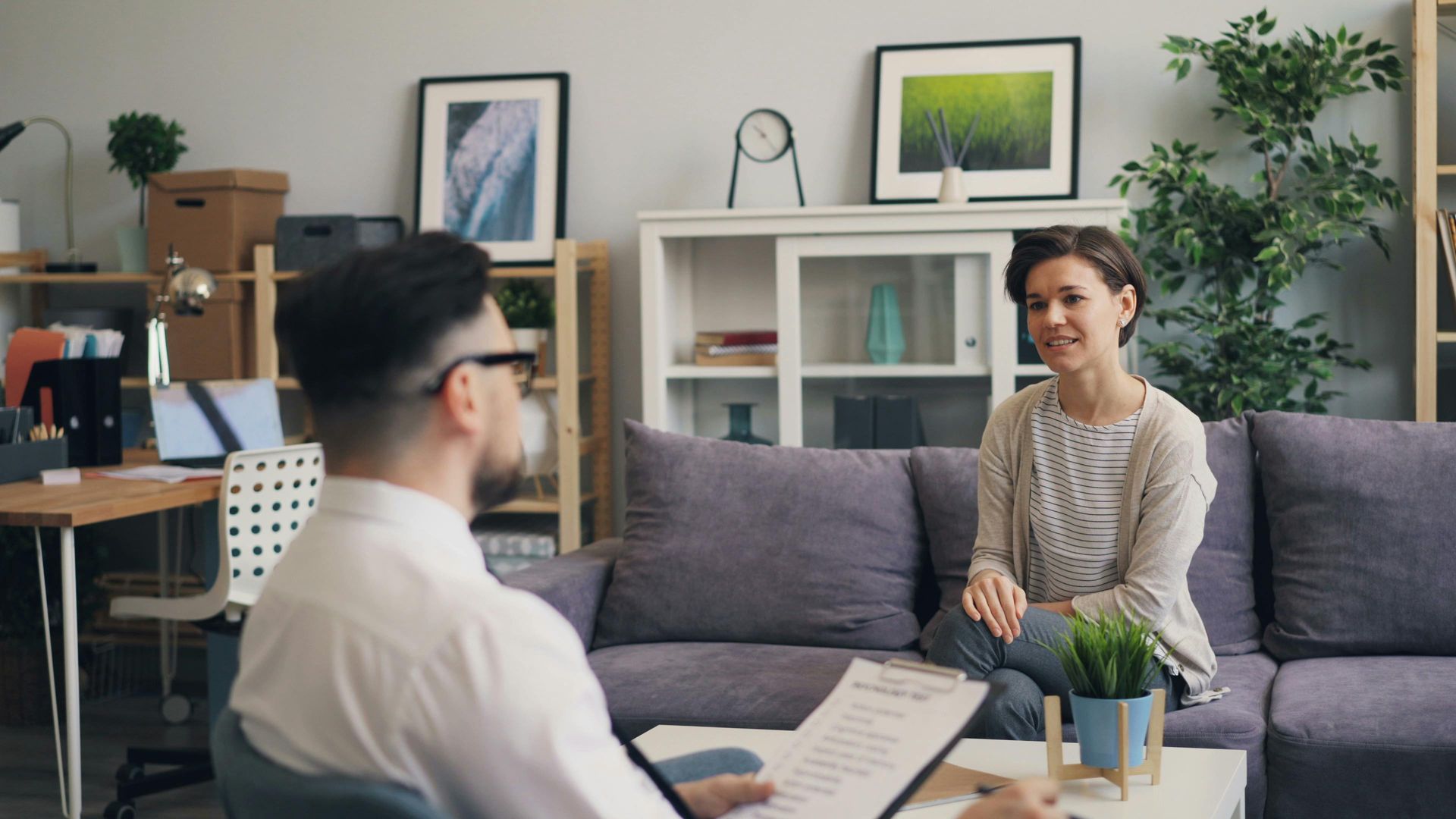 A therapist in a light room, facing a patient on a couch, taking notes.