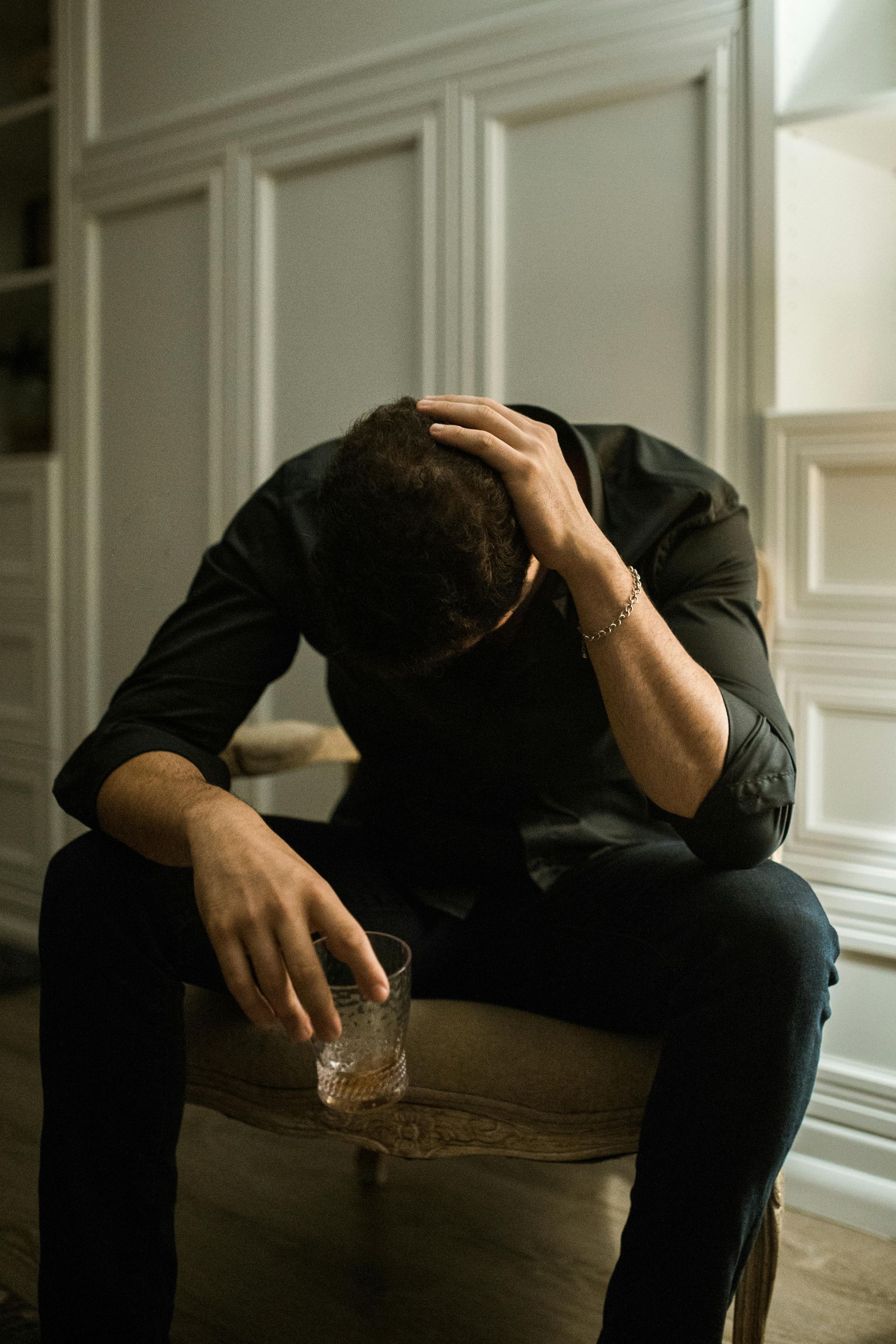Man in black shirt, head in hand, sits with drink, appearing distressed in room with ornate decor.