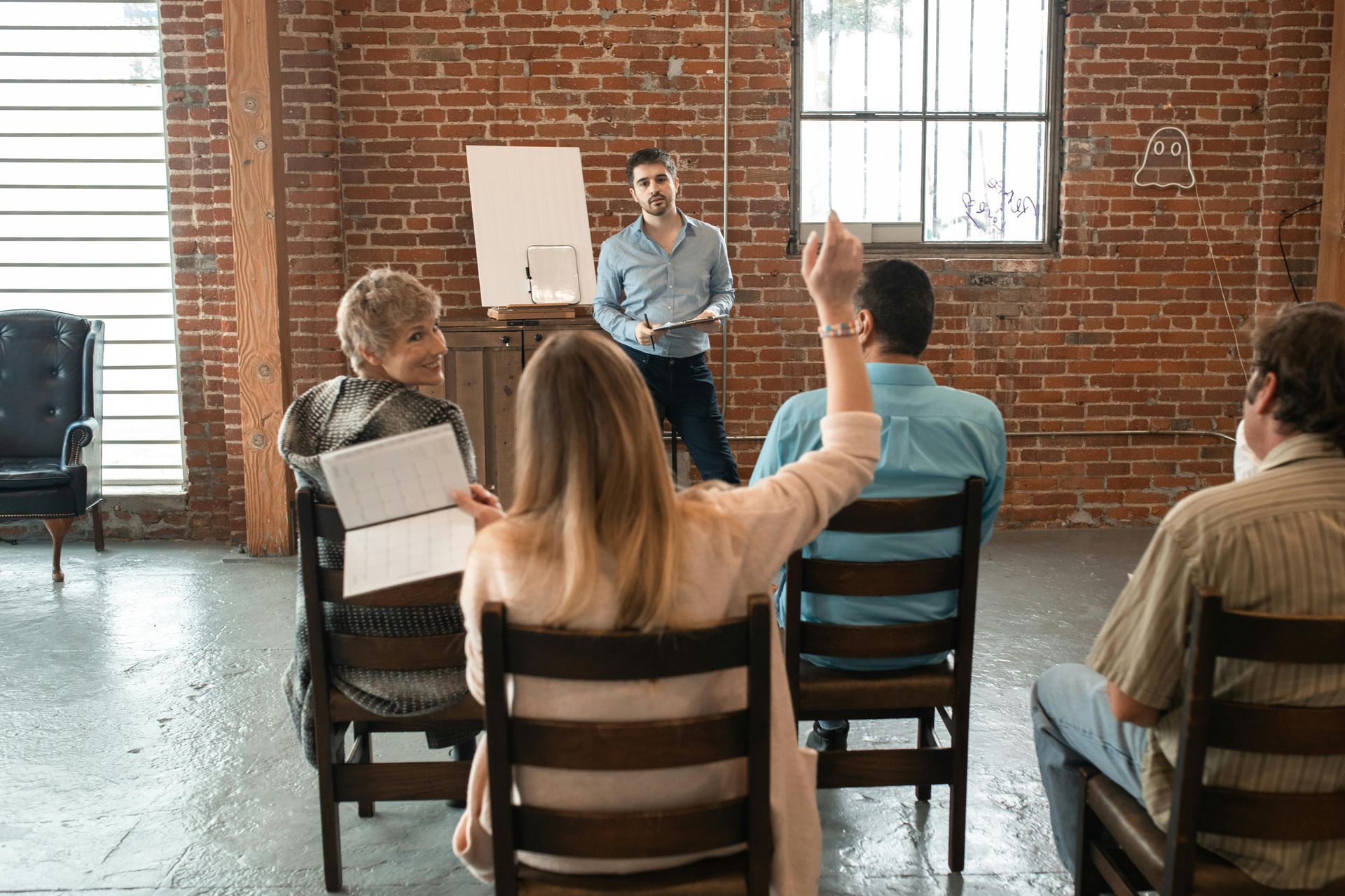 Man presents to a small group; woman raises her hand. Brick wall background, casual attire.