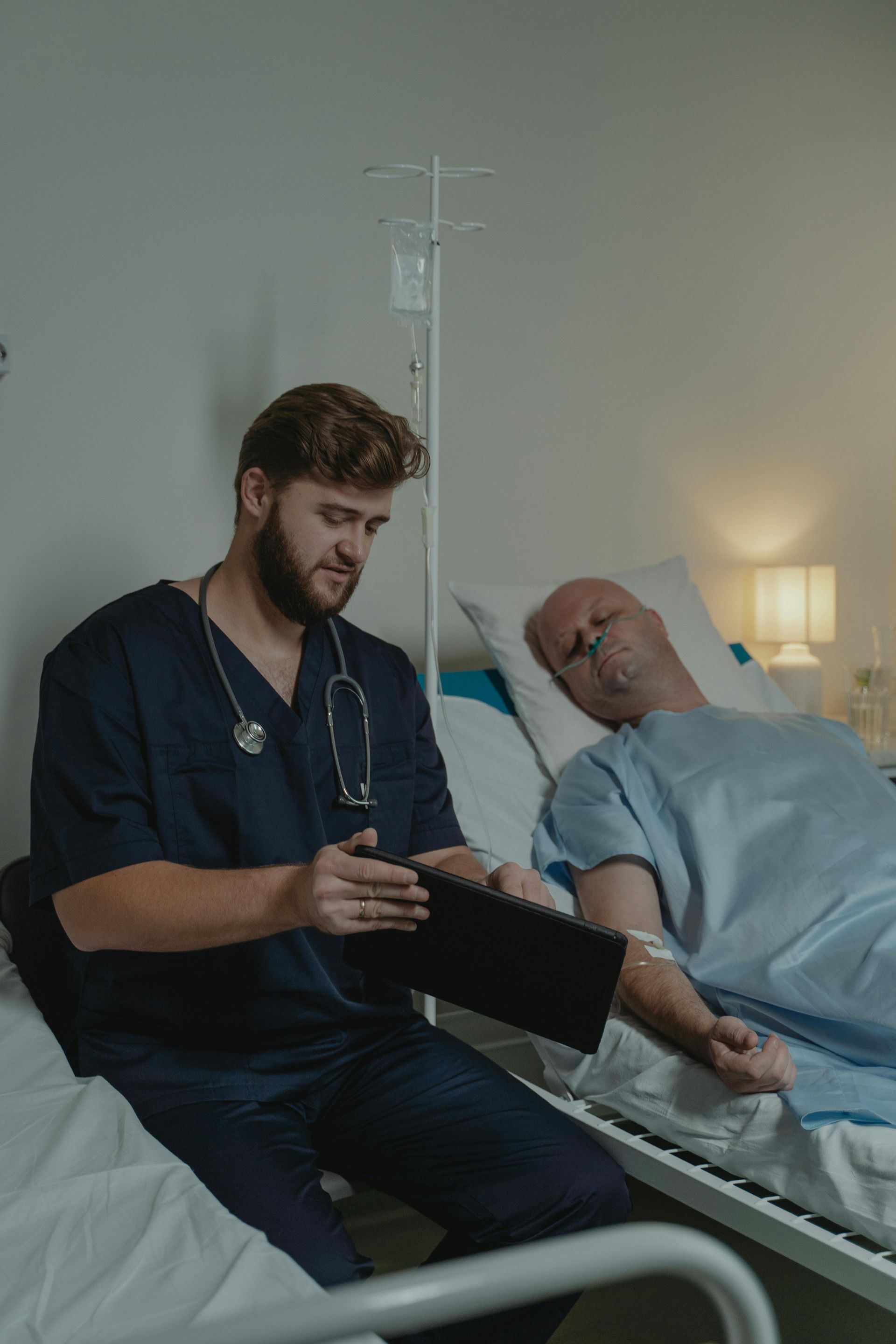 Nurse in scrubs shows tablet to patient in hospital bed with IV.