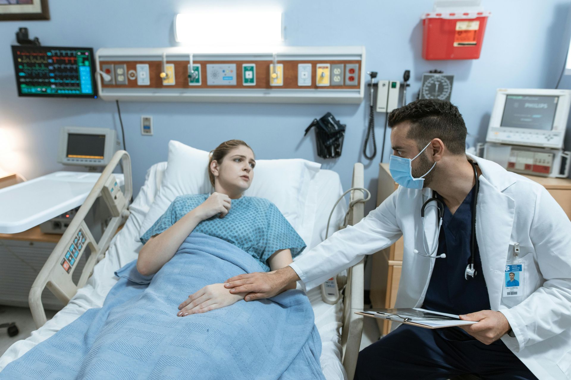 Doctor in a mask examining a patient in a hospital bed; the patient looks concerned.