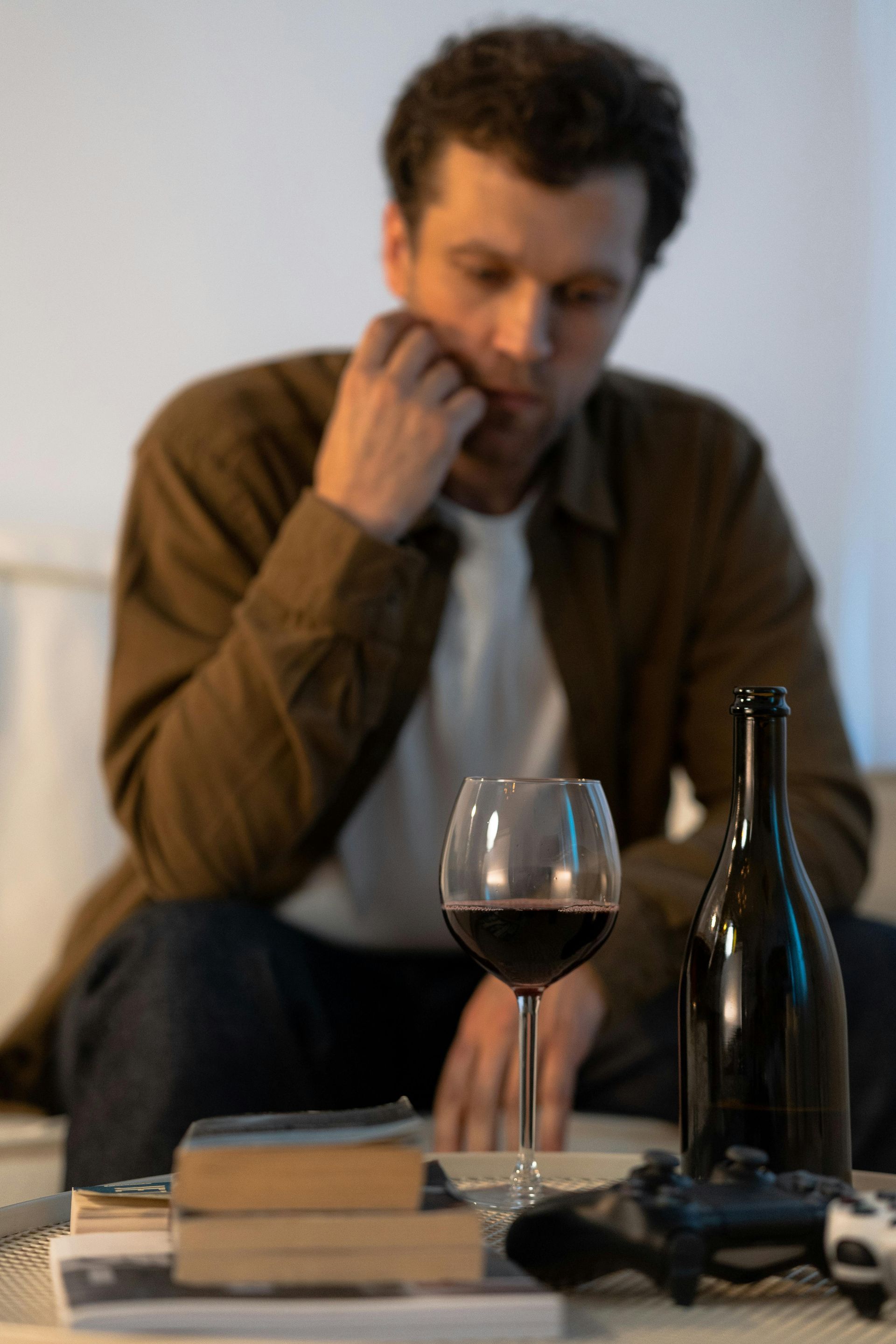 Man with wine glass, bottle, books, and game controller, appearing sad; indoors.