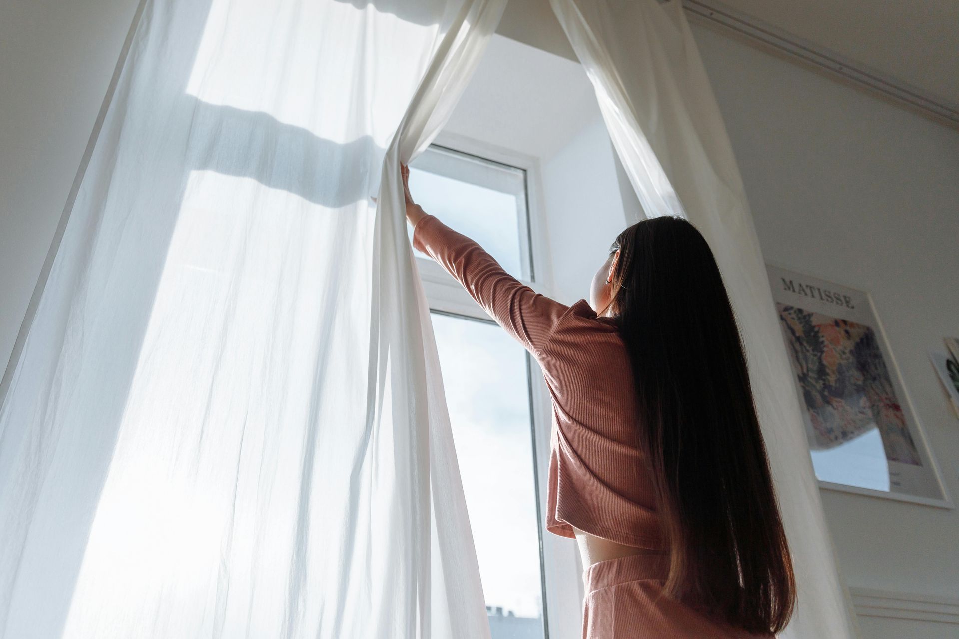 Woman with long dark hair opens white curtains, letting in sunlight.